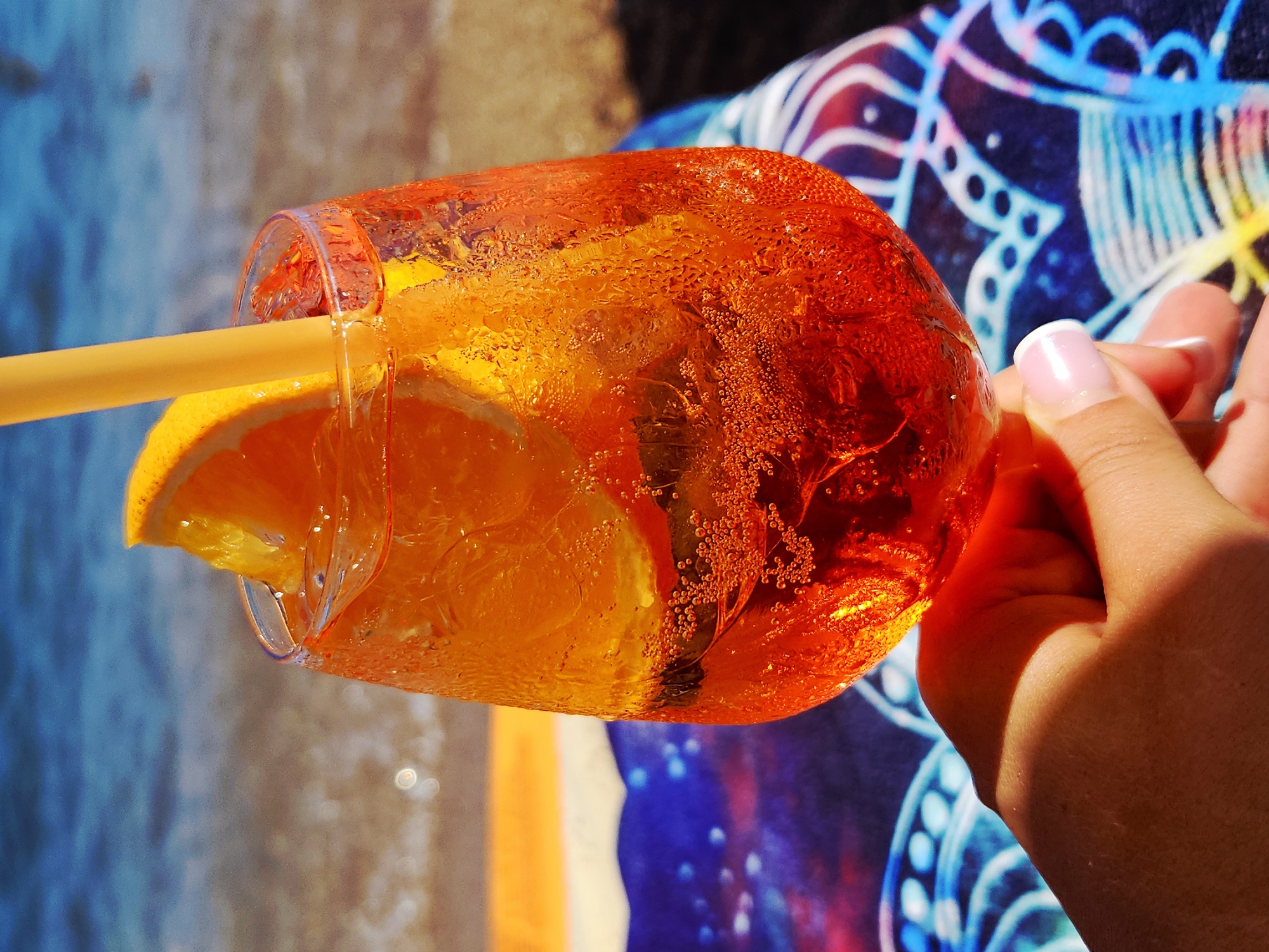 A hand holds a glass of an orange beverage with ice, a slice of orange, and a yellow straw. The glass is condensation-covered and sits on a colorful towel. In the background, the blurred image of a beach with the ocean and sand is visible. The setting appears to be a sunny outdoor location, possibly a beach.