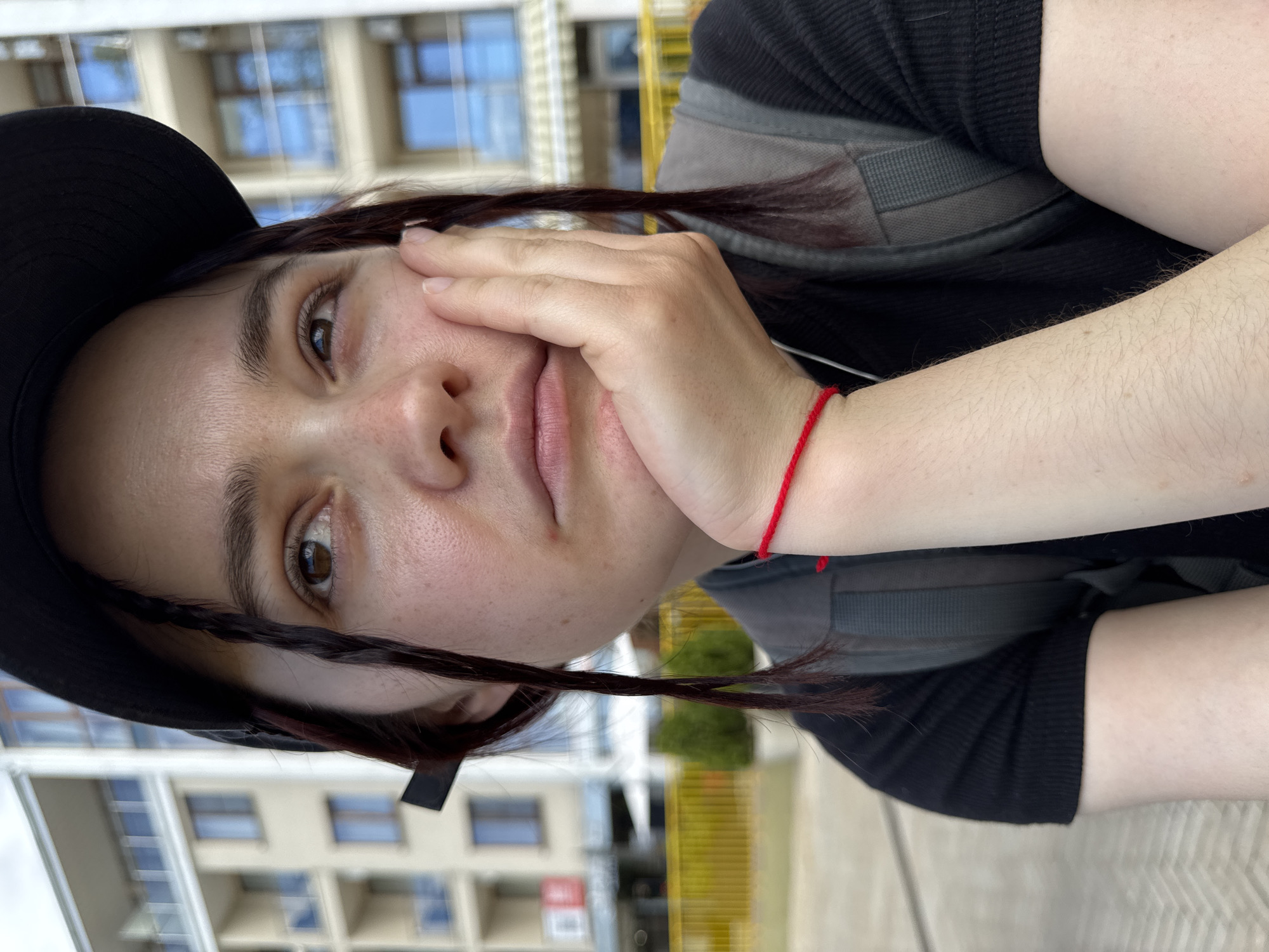 A person is posing for a close-up selfie outdoors. The subject has fair skin, dark brown hair styled with two front braids, and is wearing a black hat and a black top with a gray backpack. A red string bracelet is visible on their left wrist. The person's right hand is touching their cheek. The background features a multi-story building with windows, suggesting a location within the city. The building's design is modern, with a light-colored facade and visible window frames. The light conditions suggest it is daytime.