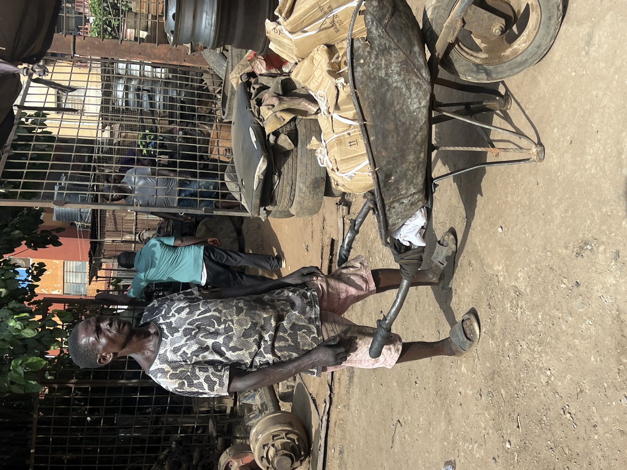 The image depicts an outdoor scene in Nkpor, Nigeria, featuring a man standing next to a heavily loaded handcart under bright daylight. The primary subject is an older man in the foreground, wearing a camouflage-patterned short-sleeved shirt, light-colored shorts or trousers, and open-toed sandals. His arms and face appear weathered, and he has a serious or weary expression, with noticeable perspiration on his forehead. He stands with his hands near the handles of a rustic, metal handcart.
The handcart itself is laden with various items, prominently featuring several cardboard boxes tied with white straps. Faint text visible on one of the boxes includes "MADE IN CHINA" and "NIGERIA." Alongside the boxes, other objects resembling vehicle parts, such as tires and metal components, are stacked. The cart has a single visible, worn wheel and robust metal handles.
Behind the man and the cart, a robust metal grate or fence partially obscures the background. Through this barrier, another man in a light teal or green shirt and dark trousers is visible, facing away from the camera. Other figures are also partially obscured behind the grate. The ground is unpaved, consisting of packed dirt and scattered gravel, suggesting a busy, practical area like a market or workshop. Green foliage from a tree or bush is visible on the left side. The strong shadows cast by the subjects and objects indicate a sunny day, likely around midday or early afternoon. The scene conveys an impression of daily labor and commerce in a bustling, semi-industrial environment.