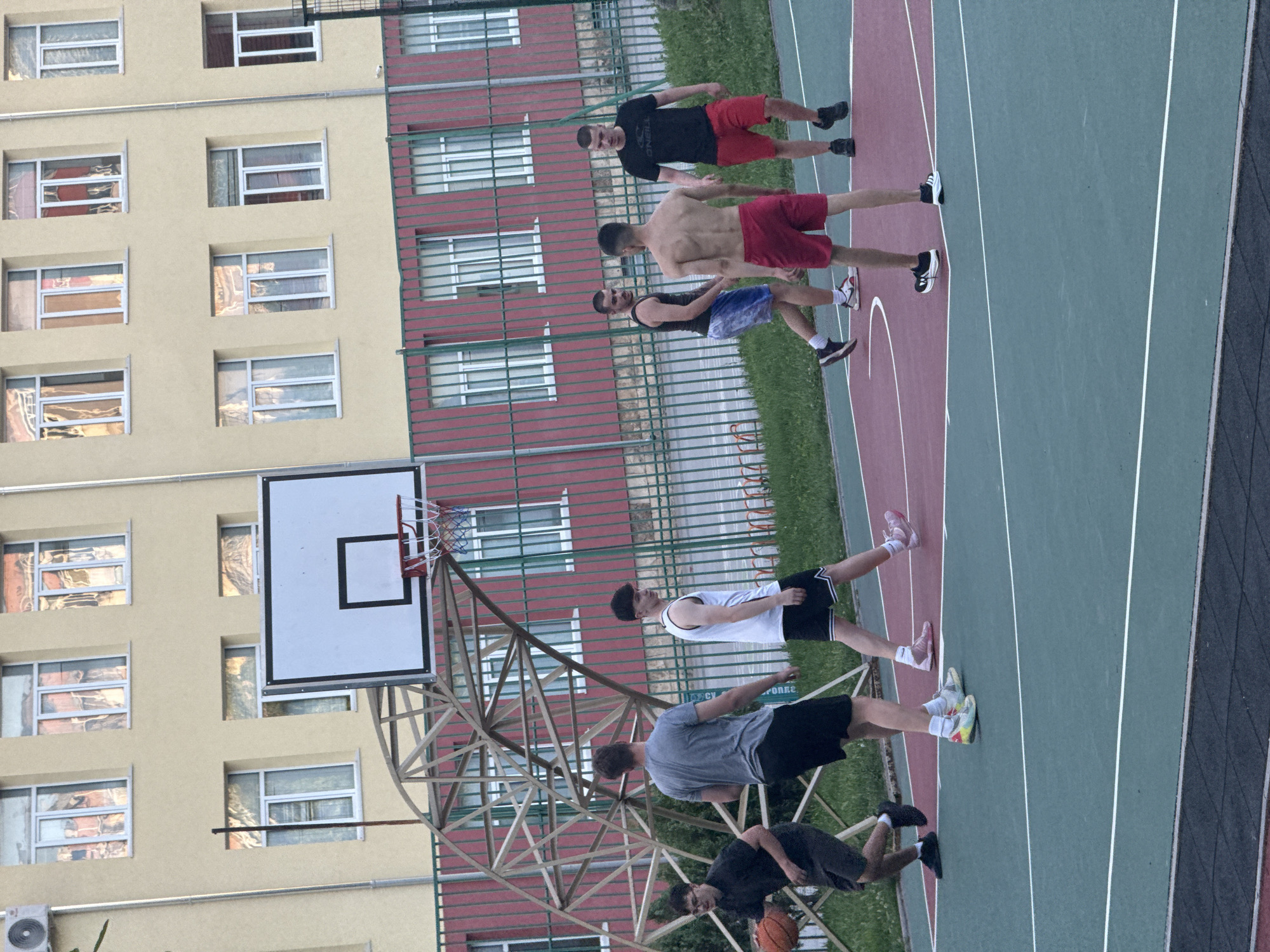 This is a medium-shot, eye-level, full-color photograph taken outdoors on a basketball court. Six young adult males are visible, engaged in a basketball game. The court has a green and red surface with white lines. In the background, a light yellow building with multiple windows stands behind a red and green fence.
The individuals are dressed in athletic wear. From left to right:
1. A male with dark hair wearing glasses, a black t-shirt, and dark shorts, dribbling a basketball with his left hand.
2. A male with dark hair wearing a gray t-shirt and black shorts, facing away from the camera.
3. A male with dark hair wearing a white tank top and black shorts with white stripes.
4. A male with short dark hair wearing a black tank top and blue and white patterned shorts.
5. A shirtless male with short dark hair wearing red shorts.
6. A male with short dark hair wearing a black t-shirt and red shorts.
The basketball hoop and backboard are prominent in the mid-ground, mounted on a wooden structure. The environment appears to be a schoolyard or public recreational area. There is no visible indication of the specific location within the city of Shumen, Bulgaria.