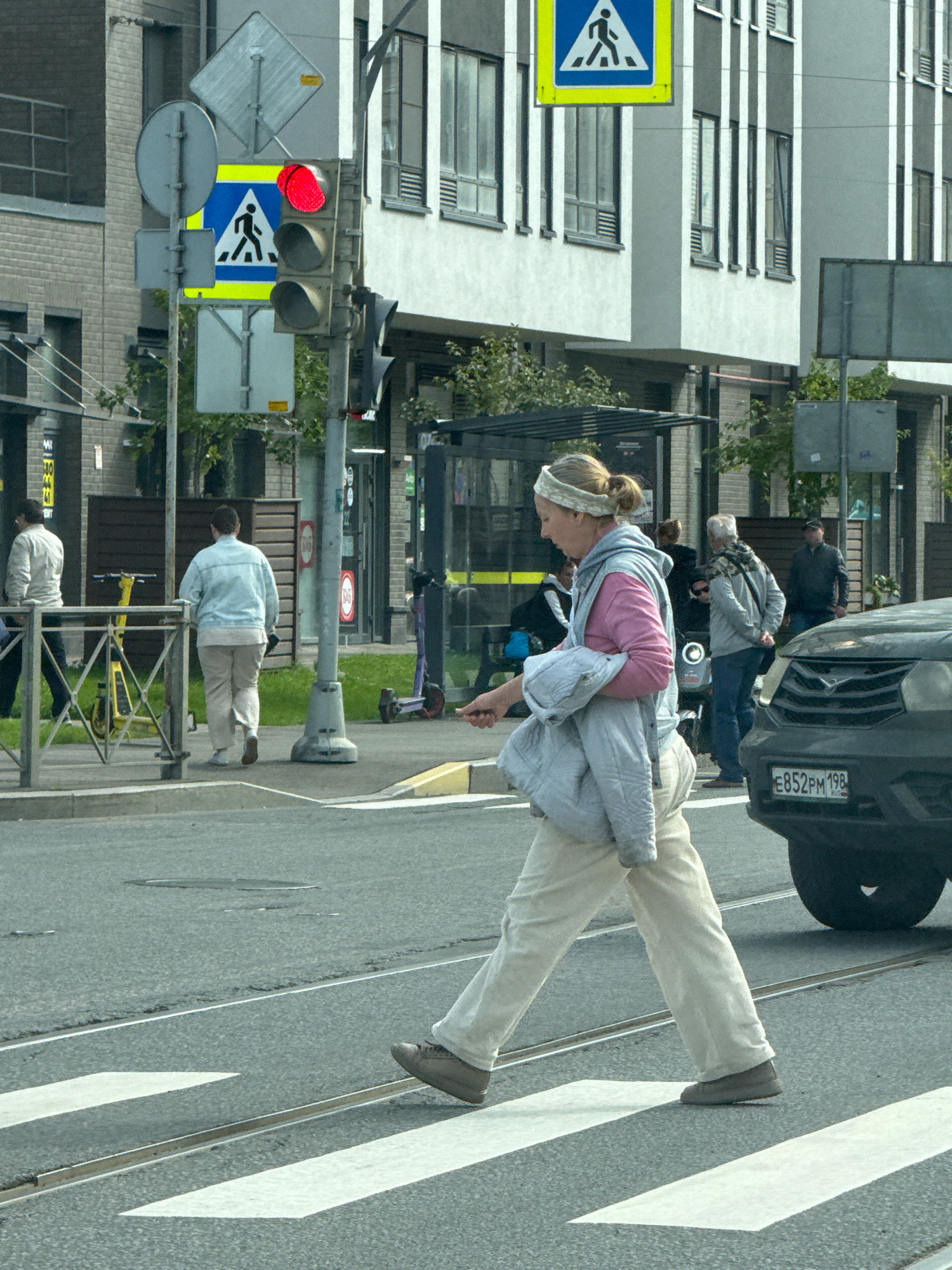 This is a street-level, medium-shot photograph taken in Saint Petersburg, Russia. In the foreground, a person is walking across a marked crosswalk from right to left. They are wearing light-colored pants, a pink hooded sweatshirt, and a grey jacket draped over their left arm. They have a light-colored headband on and their hair is pulled back. A dark-colored car is visible to the right, partially obscuring the crosswalk. In the background, a traffic light displays a red signal. Pedestrian crossing signs are mounted on a pole next to the traffic light. Further in the background, there is a modern building with a light-colored facade and dark vertical window frames, as well as a bus stop shelter. Other individuals are visible further down the street, as well as parked scooters. The street is paved, with tram tracks visible in the crosswalk. The overall environment appears to be a city street on an overcast day.