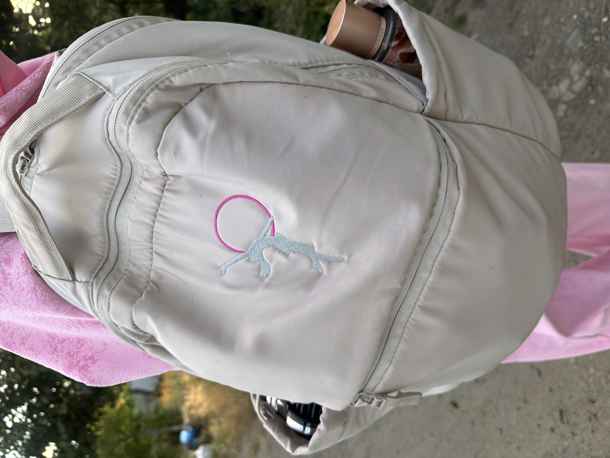 The media file shows a close-up view of a light-colored backpack, possibly being worn by a person due to the visible pink fabric on the left and right sides of the frame, suggestive of clothing. The backpack has a zippered compartment and a visible strap. On the front of the backpack, there is an embroidered design featuring a silhouette of a figure within a pink circle, with the figure appearing to be in a dynamic pose, possibly athletic or acrobatic. A metal water bottle or thermos is partially visible protruding from the top of the backpack. The background is blurred, showing hints of green foliage, suggesting an outdoor setting. The lighting indicates it could be daytime, with natural light illuminating the scene. The overall impression is casual and possibly related to an outdoor activity. No visible text is discernible in the image. The location context is identified as Dnipro, Ukraine.