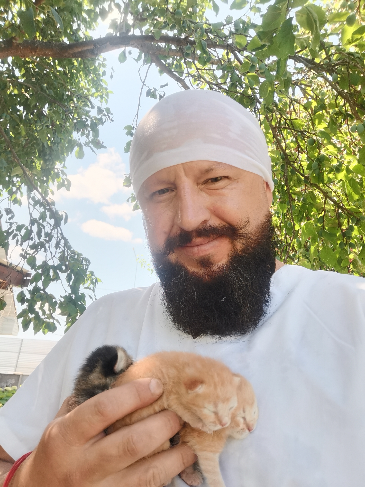 A man wearing a white headscarf and a white shirt is holding two kittens in his hands. The man has a beard and a mustache. The kittens appear to be very young. He is outdoors, with trees and foliage visible in the background, and a bright sky with clouds. The scene appears to be set in a rural or semi-rural area, possibly in the city of Semypolky, Ukraine, given the presence of trees and what looks like a fence or building in the distance. The overall lighting suggests it is daytime.