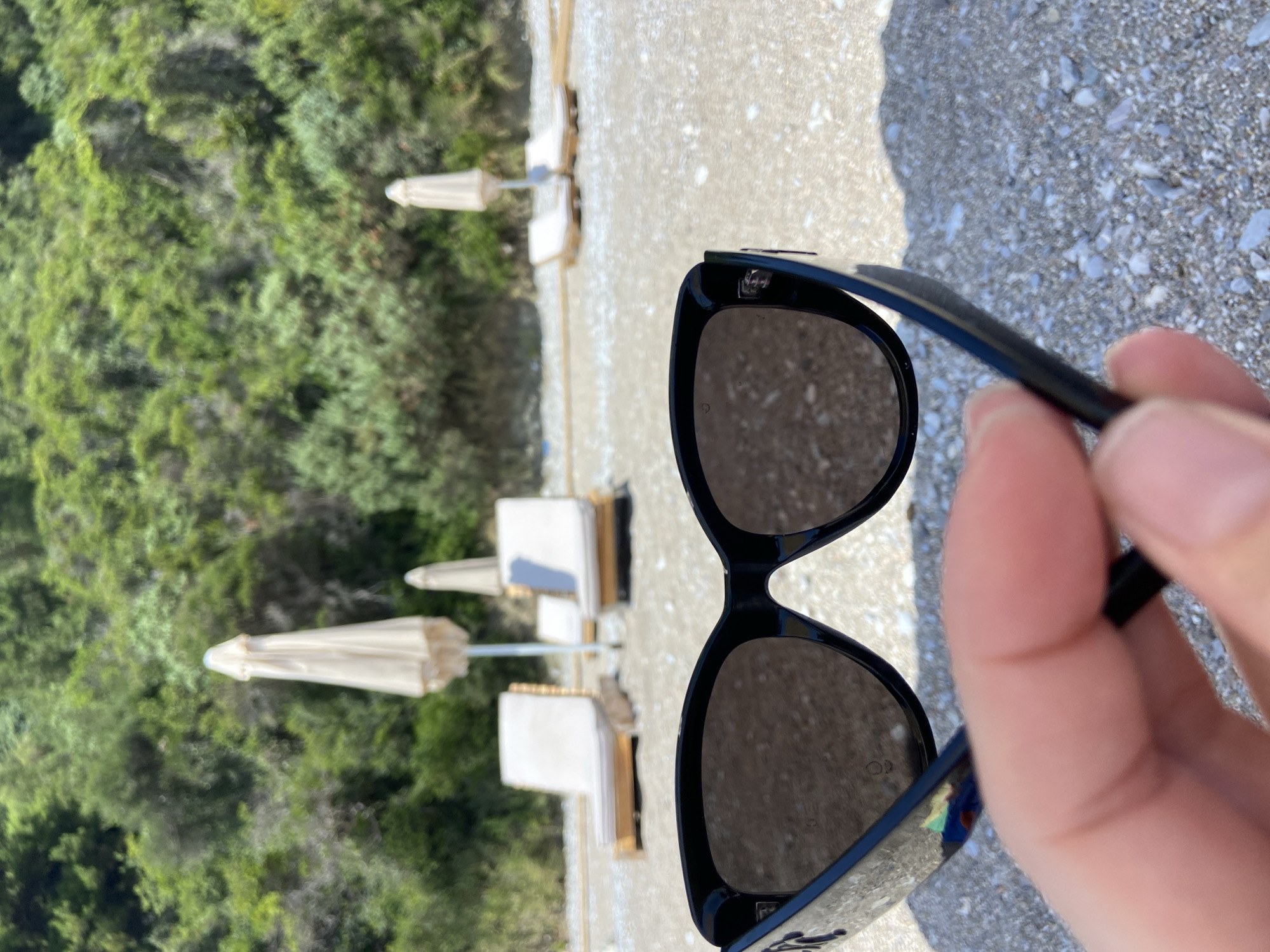 A first-person perspective shot shows a pair of black-rimmed sunglasses being held by a person's fingers in the foreground. The sunglasses are positioned such that the lenses are facing the viewer, obscuring a portion of the scene behind them. The background depicts a pebbled beach with several empty lounge chairs and closed beach umbrellas. Beyond the beach, lush green foliage covers a hillside. The scene suggests a vacation or leisure setting, possibly on a sunny day. The location is identified as Prijevor, Montenegro.