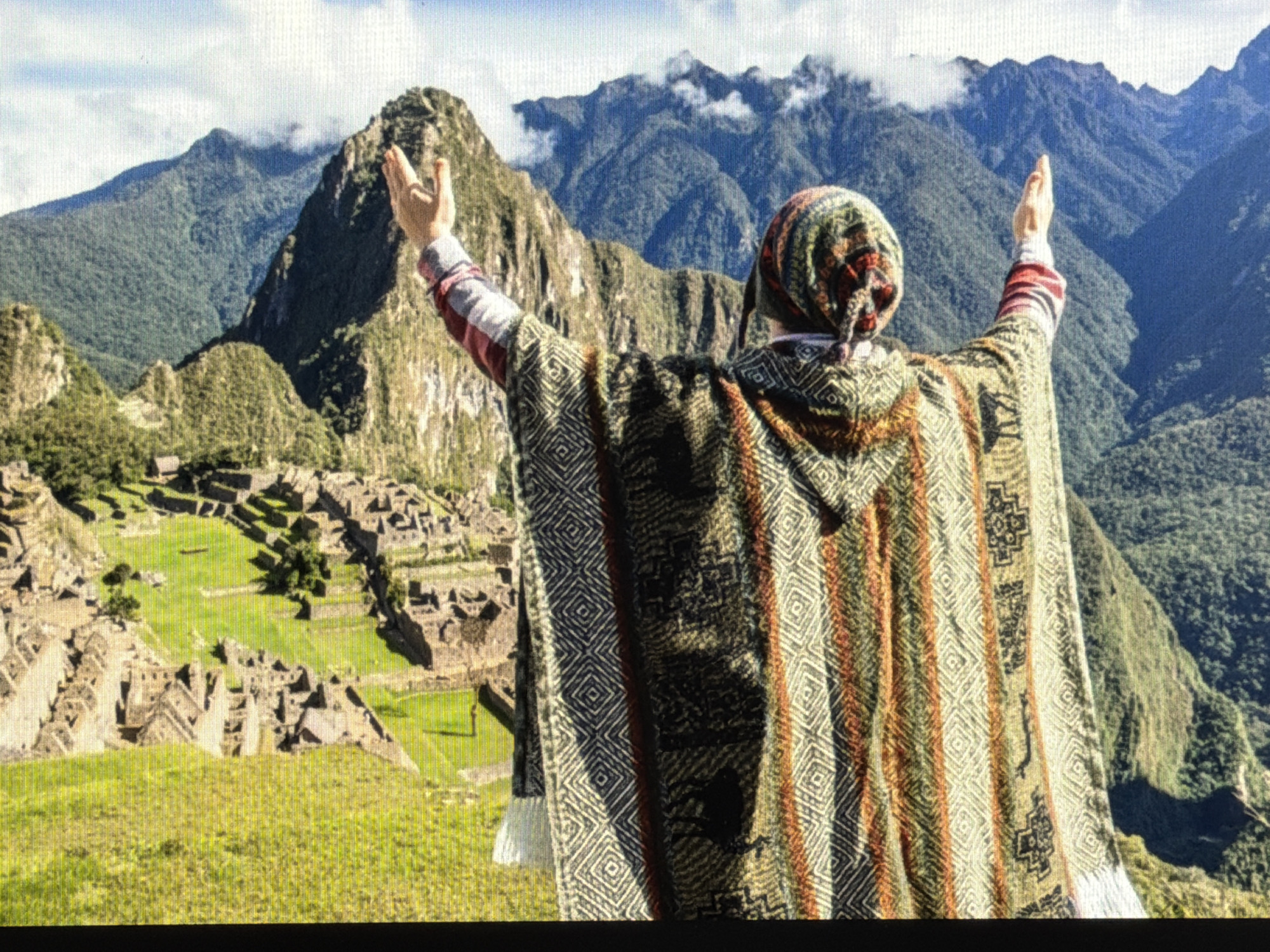 A medium, eye-level shot showcases a person with their back to the camera, arms raised, facing the ancient city of Machu Picchu in the Andes Mountains. The individual is wearing a hooded, patterned poncho with white fringe and a similarly patterned head covering. Machu Picchu is visible on a green, grassy plateau, with intricate stone structures and ruins. Behind Machu Picchu, towering, green mountains rise into the sky, partially obscured by clouds. The environment consists of lush vegetation and clear blue skies. The action captured is the person raising their hands, seemingly in reverence or celebration of the location. The interaction is between the person and the landscape of Machu Picchu.