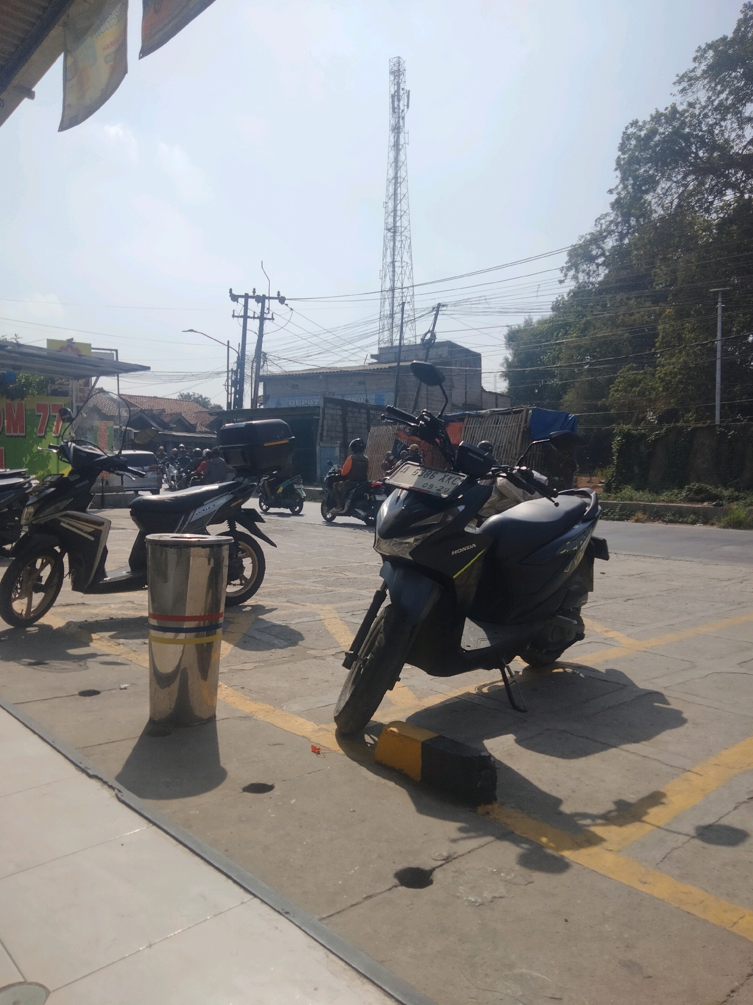 The image captures an outdoor scene with several parked motorcycles in what appears to be a parking lot adjacent to a street. In the foreground, a silver cylindrical trash can with red and yellow bands is prominent, casting a distinct shadow. Two black scooters are parked to the left of the trash can, and another black scooter with a visible license plate (A 6386 XKC) is positioned in the center right of the frame. The scooter in the center right also has a black storage box on the rear. Behind the parked scooters, other motorcycles are visible, some with riders.
The environment suggests a bright, sunny day with a clear sky, though a tall communication tower is visible in the distance. Power lines and trees are also present in the background, indicating an urban or semi-urban setting. The ground is paved with concrete and marked with yellow parking lines. A building with signage that includes "DEPOT" is visible in the mid-ground. The overall impression is that of a public area, possibly near shops or a service establishment, in Pisangan Jaya, Indonesia.