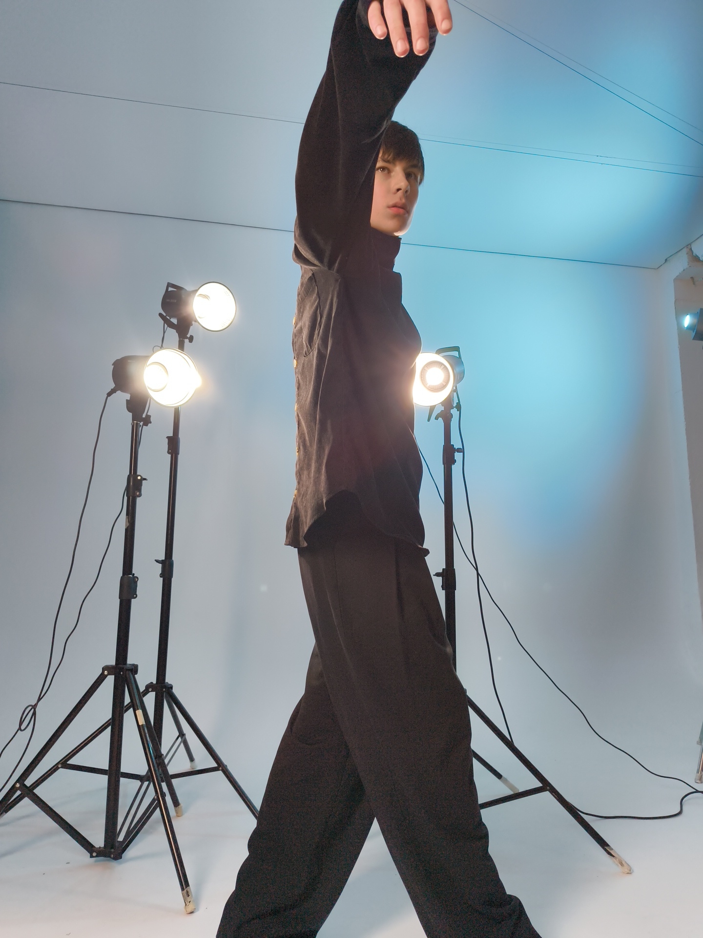 A person in a black turtleneck, black jacket, and black pants is posing in what appears to be a photo studio. The person's left arm is raised, and they are looking off to the side. Three studio lights on stands are visible, providing illumination. The background is a neutral gray or blue color. The location appears to be a studio based on the presence of the studio lights and simple backdrop. There is no evidence that the image was taken in Chaikovsky, Russia.