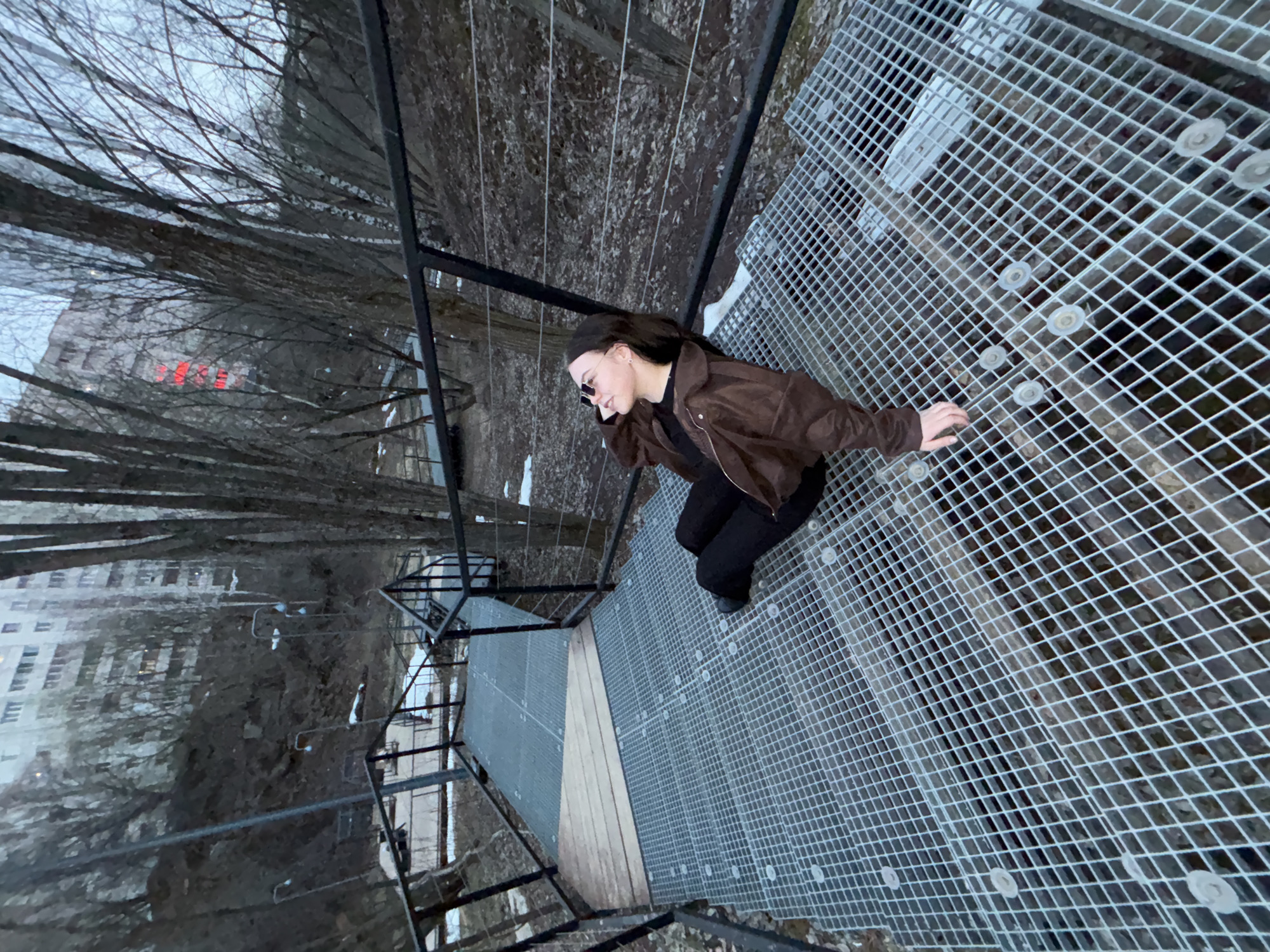 A medium shot from above depicts a young woman crouching on a metal mesh walkway. She wears a brown jacket, black pants, and sunglasses, and her head is slightly bowed. The walkway is elevated and part of a metal structure that appears to be a pedestrian bridge or observation platform. Beyond the structure, trees and buildings are visible, suggesting an urban setting. The light suggests overcast conditions. The walkway is located in Perm, Russia.