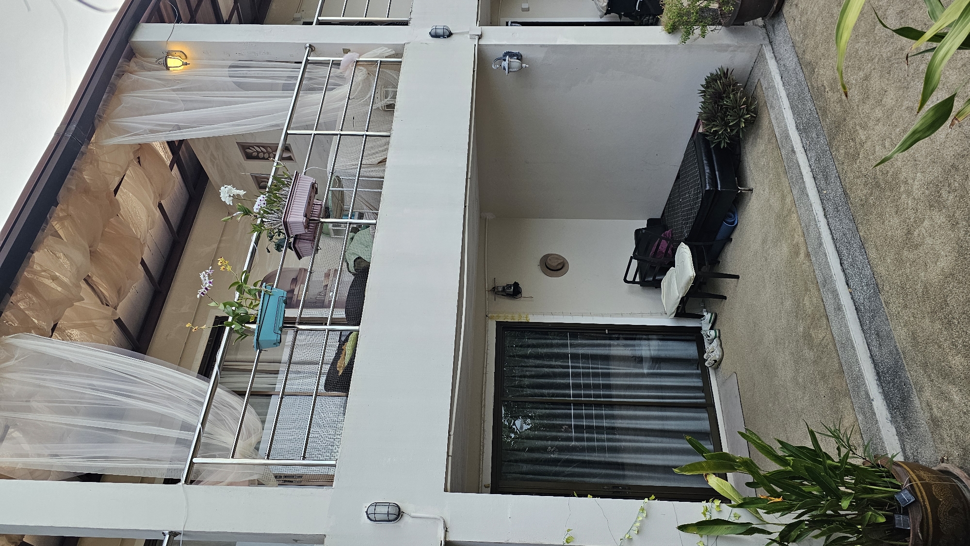 This is a street-level, exterior, medium shot of a building's facade in Baan Chaweng, Thailand. The building appears to be two stories tall with a balcony on the upper level and a covered patio on the lower level. The balcony is adorned with plants in blue and pink planters, white sheer curtains, and a stainless steel railing. On the lower level, there is a sliding glass door with vertical blinds, a doormat, a pair of sneakers, a small chair with a white cushion, a black daybed, and a rolled-up blue mat. A brown hat is hanging on the wall. The patio floor is concrete. To the left of the patio, there is a large potted plant with green leaves. The building's exterior walls are white. The lighting suggests it is either early morning or late afternoon. There are no people visible in the image.