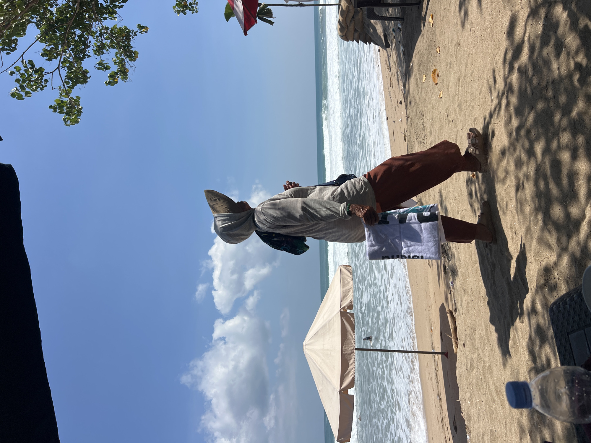 A person wearing a headscarf, a light-colored long-sleeved shirt, and dark orange pants stands on a sandy beach in Kuta, Indonesia, facing right in profile. They are holding a white towel with dark lettering. In the background, the ocean has moderate waves, and there is a large beige umbrella on the left and a red umbrella on the right, near sandbags. The sky is blue with scattered white clouds. The scene appears to be in the afternoon, given the shadows.