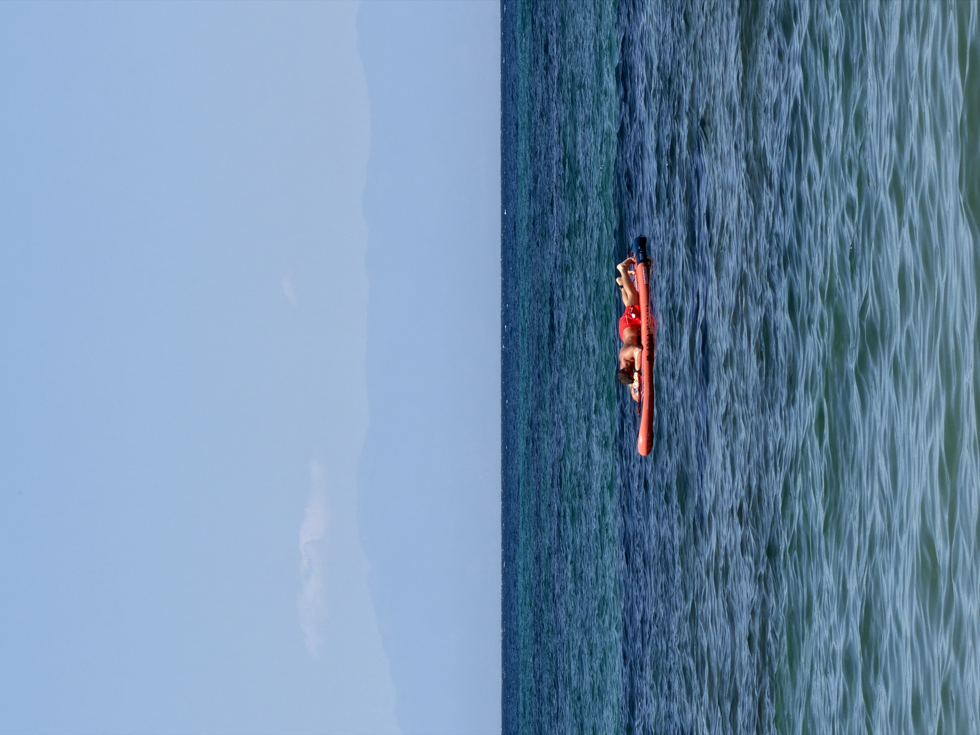 A person is lying on a red inflatable raft in the sea. The raft has two blue handles. The person is wearing red shorts. The sea is a deep blue color with small waves. The sky is a light blue color with some clouds. The image is taken from above, giving a clear view of the person and the sea. The scene suggests a sunny day at the beach. There is no visible text in the image. The photo was taken in Nea Iraklia, Greece.