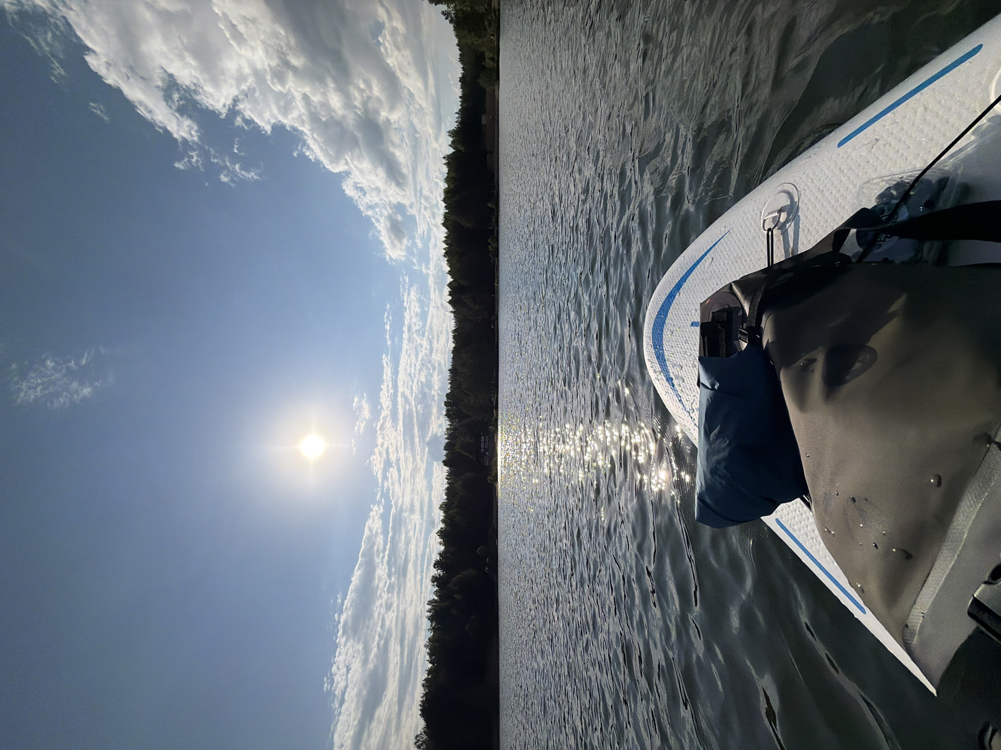 The image shows a view from a paddleboard on a body of water, likely a lake or river. The paddleboard is white with blue accents and has a dark-colored bag attached to it. The water has visible ripples and reflects sunlight, creating shimmering highlights. In the background, there is a treeline indicating the shoreline. The sky is blue with scattered white clouds and the sun is visible. While the prompt mentions Selychka, Russia, there is no information in the image that confirms that this is the location.
