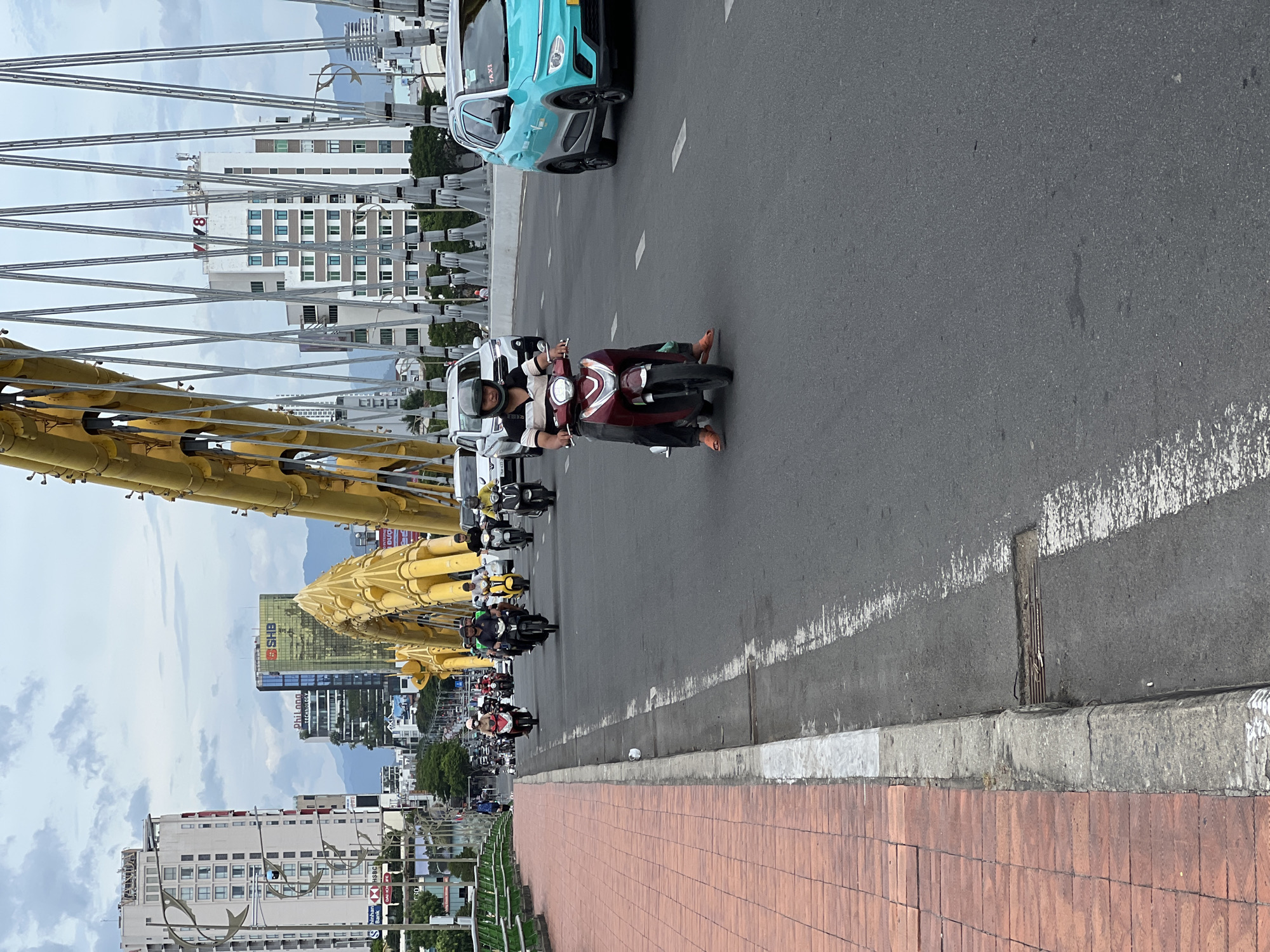 This image depicts a daytime scene on the Dragon Bridge (Cầu Rồng) in Đà Nẵng, Vietnam. The bridge features a prominent yellow, dragon-shaped superstructure with numerous suspension cables arching over the roadway.
In the immediate foreground, a man wearing a black and white striped polo shirt, black trousers, a black helmet, and orange sandals operates a dark red scooter. He is positioned towards the center of the right lane and gazes forward. To his right, a light blue-green VinFast VF e34 electric SUV, identifiable as a "TAXI" by pink lettering on its roof, travels in an adjacent lane. Behind the foreground scooter, numerous other motorbikes and several white cars are observed moving across the bridge.
On the far left, a terracotta-tiled pedestrian sidewalk is bordered by a green metal railing. A single pedestrian is visible walking in the distance on this sidewalk. The asphalt roadway displays faded white dashed lane markings and a concrete curb. A small rectangular drain opening with metallic grates and some debris is located near the curb.
The urban background reveals multiple high-rise buildings, including the gold-facaded SHB bank, Phi Long Technology, HSBC, and VTV8, all under an overcast sky with scattered clouds. Mountains are discernible in the distant background. The overall scene indicates active traffic flow on a major urban bridge.