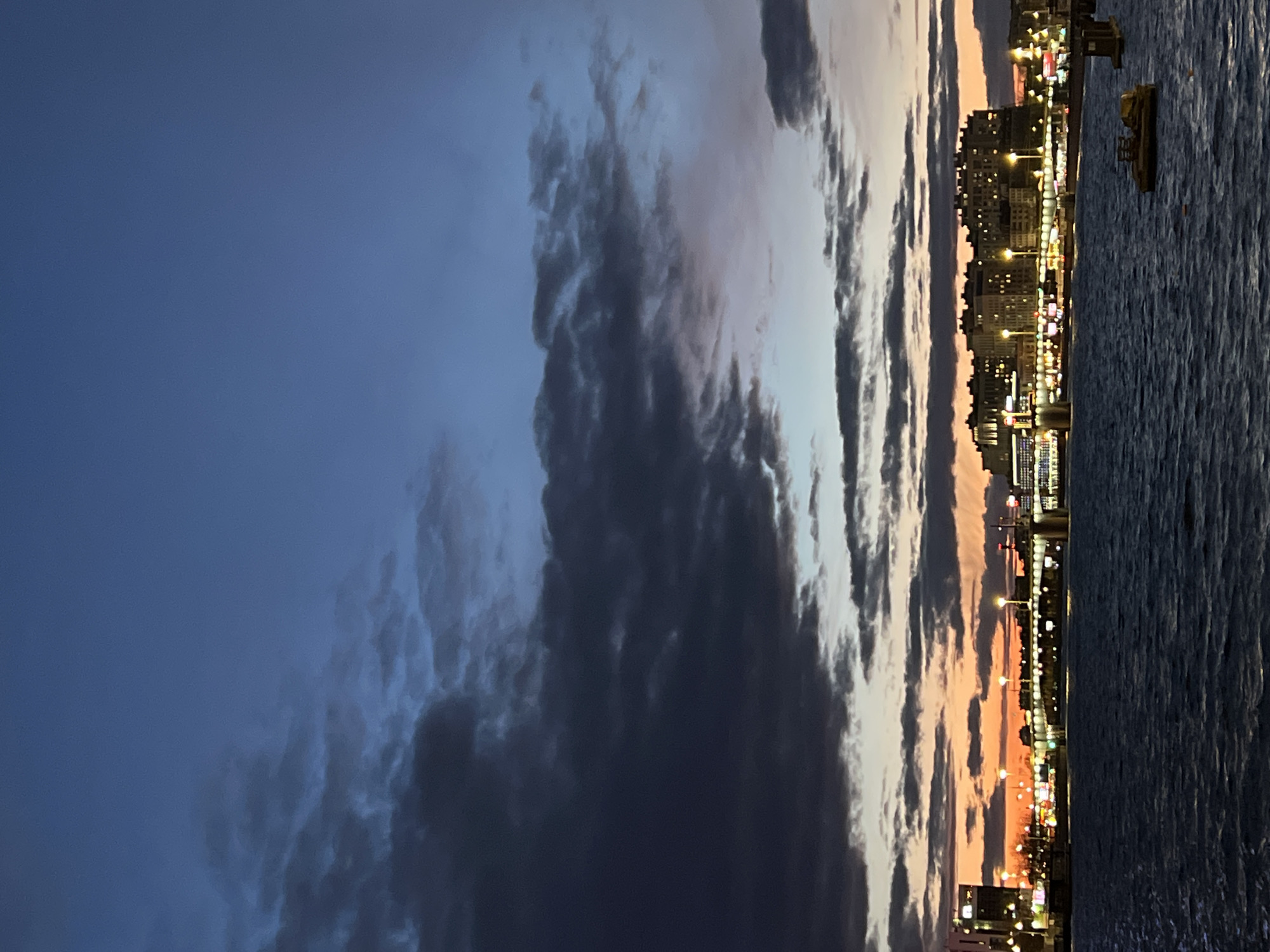 This media file captures a captivating dusk or nighttime scene of a waterfront cityscape, likely in Saint Petersburg, Russia. The image is composed horizontally, with a dramatic sky filling the upper portion, a brightly lit city skyline in the middle, and a dark body of water in the foreground.
The sky exhibits a striking gradient of colors, transitioning from a deep, somber blue on the left to lighter, warmer hues of orange and pink near the horizon on the right, indicating either sunset or the lingering afterglow of daylight. Dense, dark, and textured clouds stretch across the sky, creating a sense of depth and movement. Some clouds appear as elongated, parallel formations, contrasting with the more amorphous shapes in the darker sky.
Below the sky, the city skyline comes alive with a multitude of illuminated buildings. These structures, appearing as dense urban development, feature numerous glowing windows and streetlights that cast bright points of light across the scene. The lights along the waterfront create a continuous, shimmering line, suggesting a bustling promenade or embankment. The architecture appears typical of a European city.
In the foreground, a vast expanse of dark water reflects some of the city lights faintly, though the surface remains largely shadowed and rippled. A prominent yellow object, possibly a boat or a fixed structure, is visible on the water to the right, near the city edge.
The overall impression is one of urban tranquility and dramatic natural beauty, capturing the transition from day to night in a vibrant city. There are no people or discernible text visible in the image.
