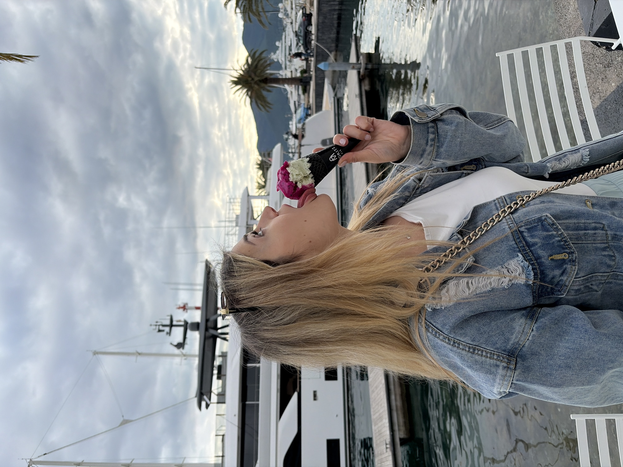 A young woman is eating ice cream at a marina in Tivat, Montenegro. She has long blonde hair and is wearing a denim jacket. She is seated next to the water, with yachts visible in the background. The ice cream is in a black cone and appears to be multi-flavored. The weather is overcast.