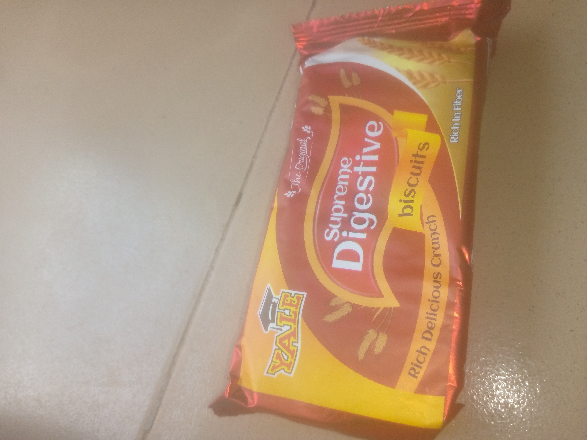 A package of "Supreme Digestive Biscuits" is displayed on a beige tiled surface. The package is mostly orange and yellow, with red accents. The brand name "YALE" is visible in the lower left corner of the package. Text on the package includes "The Original," "Supreme Digestive," "biscuits," "Rich Delicious Crunch," and "Rich In Fiber." The lighting in the image is somewhat diffused, creating a soft glow.