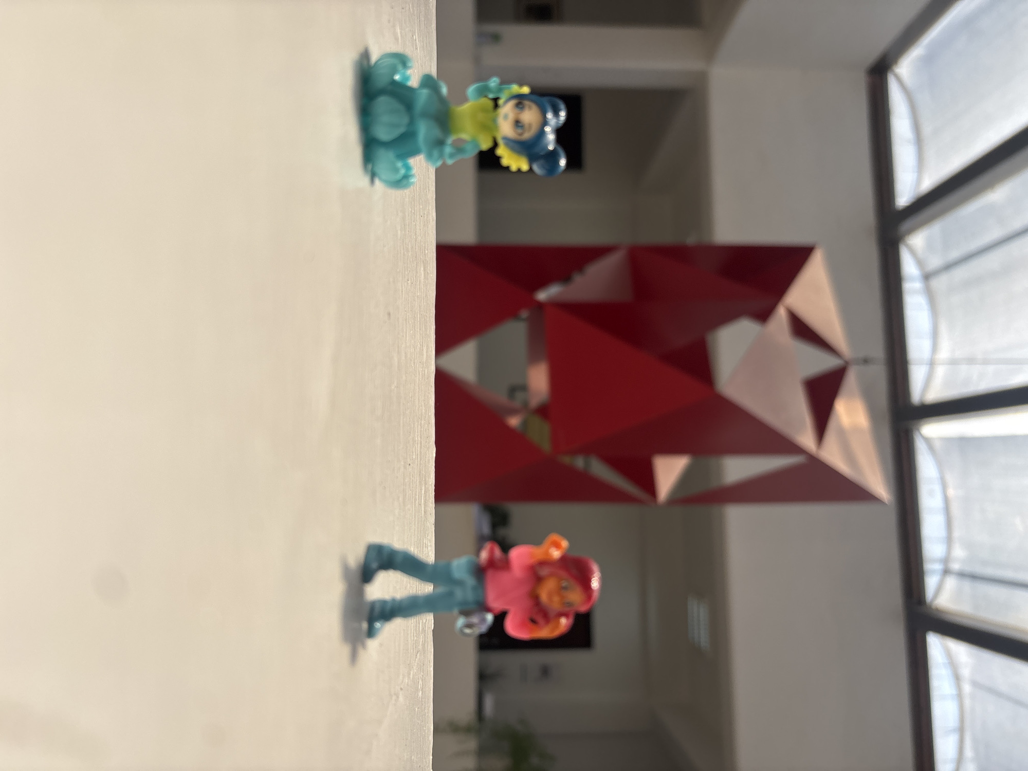 On a white ledge are two small plastic figurines, one with blue hair and a blue ruffled dress, and another with reddish hair and a pink shirt. Behind them is a red geometric sculpture and a window. The location is in San Andrés Cholula, Mexico.