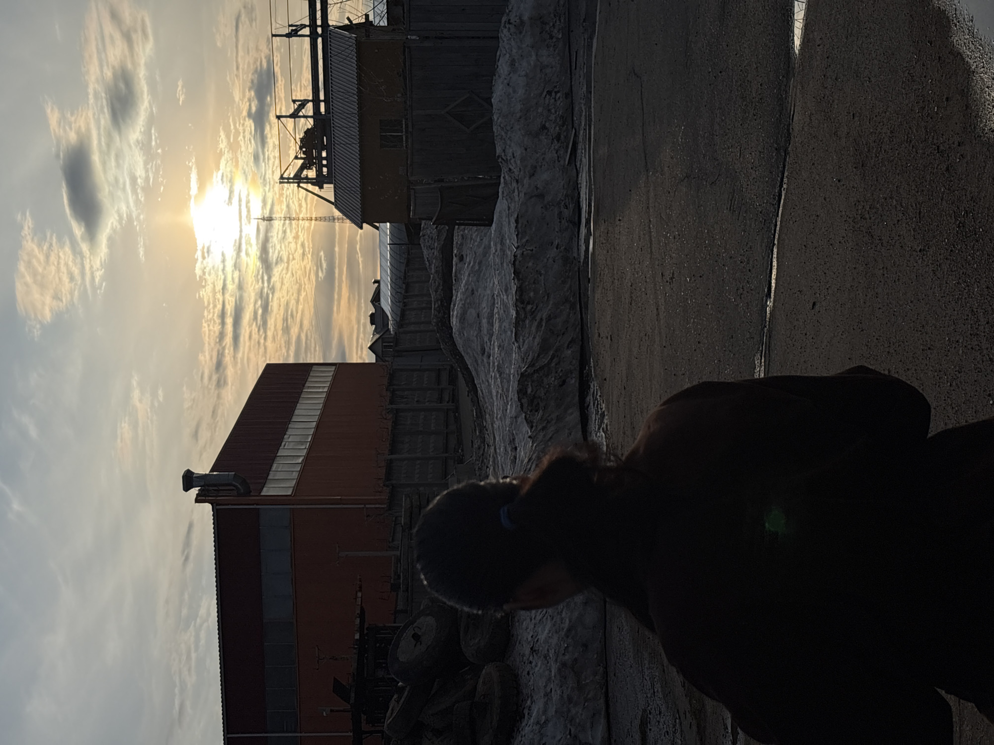 The image shows a person standing on a paved surface in what appears to be an industrial or semi-industrial area of Chaikovsky, Russia. The sun is low in the sky, obscured by clouds and illuminating the scene with a warm glow. In the background, there are buildings, possibly warehouses or factories, with visible architectural elements. Snow is present, indicating a cold climate. There is some sort of structure of what seems to be an old shed on the upper right side of the image. A tall metallic structure is also visible in the center to left center of the image behind the building.