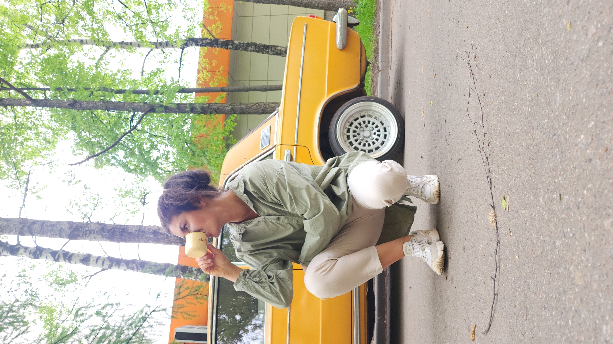A woman is squatting and drinking from a small, light-yellow cup. She wears a light-green, button-down shirt, light-tan pants, and white sneakers. Behind her is a yellow vintage car, parked in front of a building with orange and white sections. Trees with light-green foliage are in the background. The location is likely Moscow, Russia, given the style of the car. A thin, dried branch lies on the asphalt beside her.