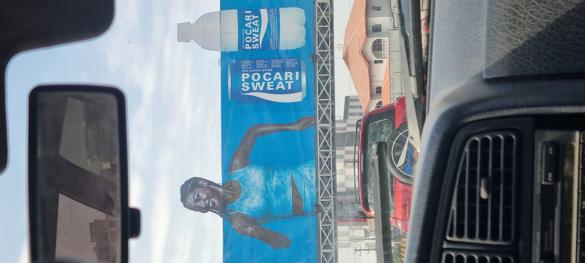 The image depicts a street view from inside a vehicle, looking towards a large billboard. The billboard features an advertisement for Pocari Sweat, an ion supply drink. The advertisement prominently displays a smiling woman in athletic wear running, alongside images of a Pocari Sweat can and bottle. The billboard is positioned above a road, with a red SUV visible in traffic below it. Beyond the road, residential buildings with orange tiled roofs are visible, characteristic of urban or suburban areas. The sky is cloudy. The interior of the car shows a dashboard with air vents and a radio. The scene appears to be in Ikoyi, Nigeria, based on the context provided.