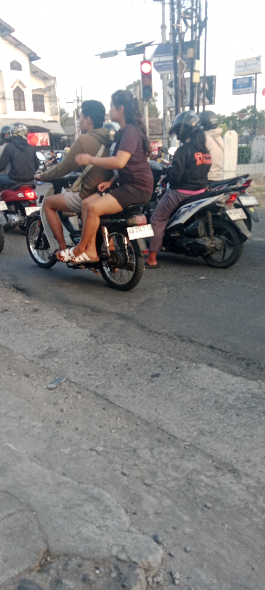 In Maguwoharjo, Indonesia, an outdoor daytime scene captures a traffic intersection with multiple motorcycles. A red traffic light indicates a stop. On the left, a motorcyclist in a black jacket with a helmet is partially visible. Beside them, a couple is riding a black motorcycle. The man is wearing a brown hoodie and grey shorts, and the woman is in a dark purple t-shirt and dark shorts. The motorcycle has a license plate that reads "AD 3147 TB." Behind them, another motorcycle carries two individuals, one in a black jacket with a logo and the other in a light-colored jacket. The road surface is paved and appears worn. Buildings and power lines are visible in the background, suggesting an urban environment. The sky is bright, indicating daytime.