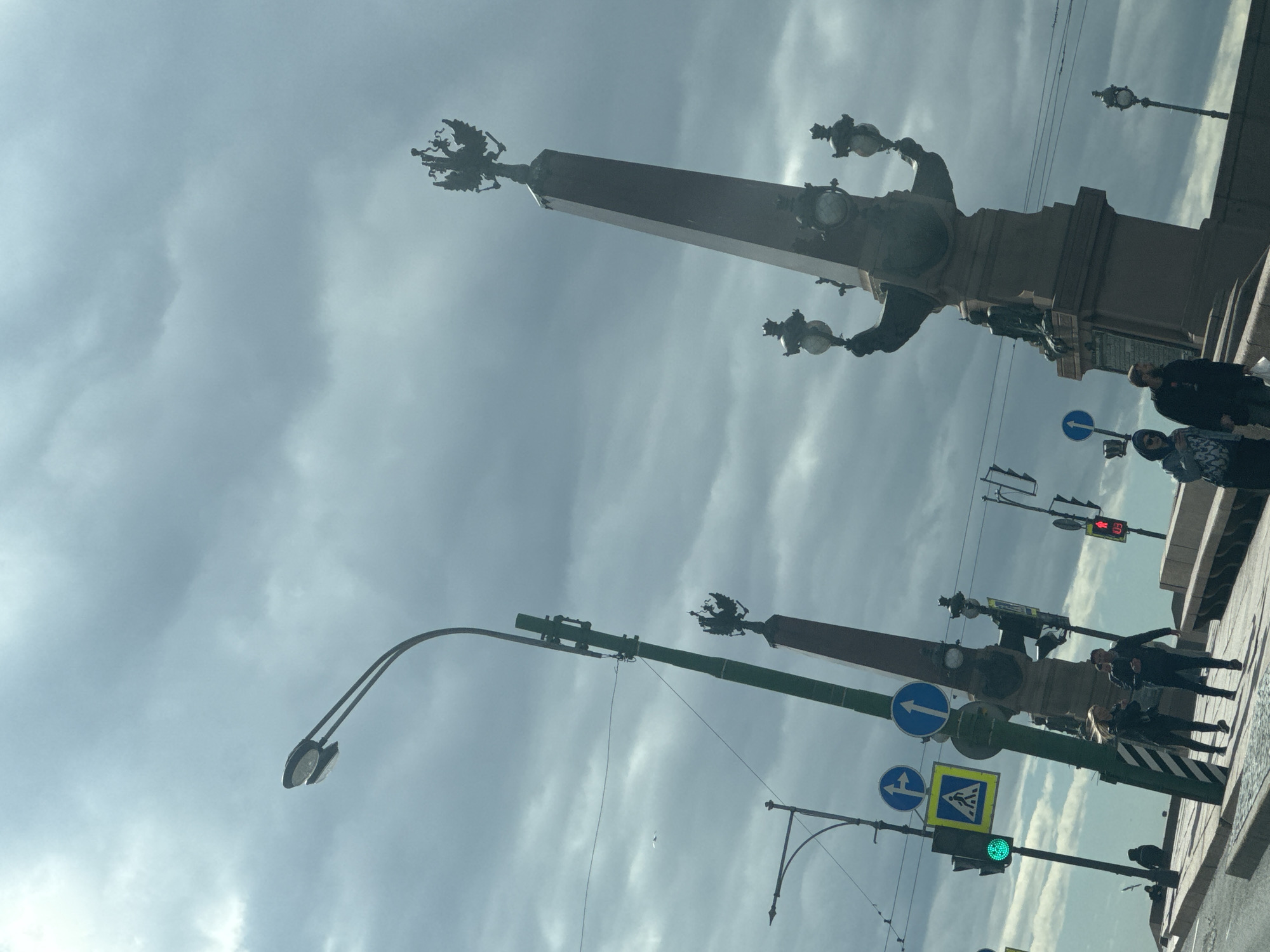The image shows a view of the Rostral Columns on the Vasilievsky Island side of the Exchange Bridge in Saint Petersburg, Russia. The columns are topped with rostral ornaments and stand against a backdrop of a cloudy sky. People are walking along the sidewalk next to the bridge. Streetlights and traffic signals are also visible. One traffic light is green.