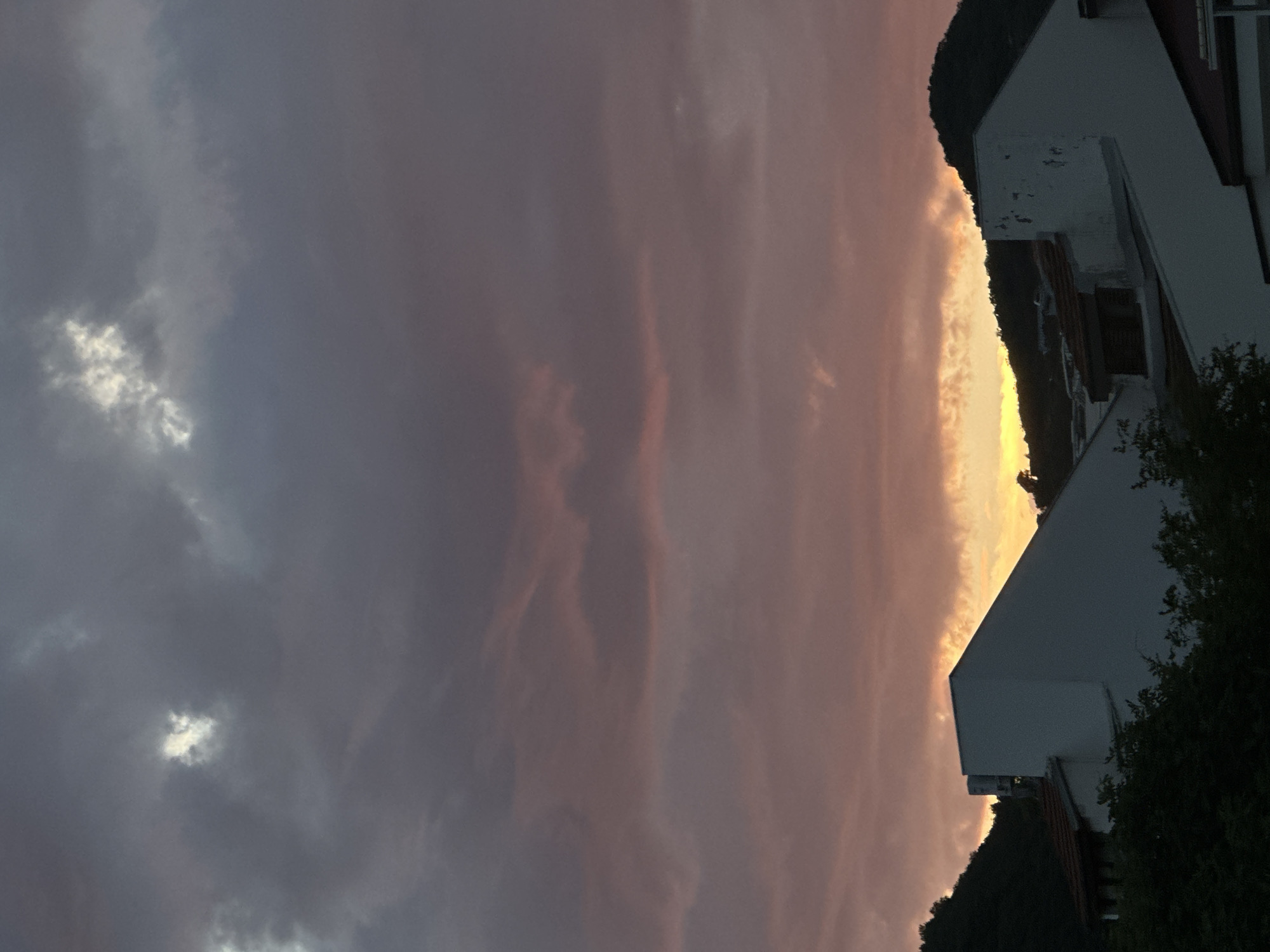 An outdoor scene captures a twilight sky over Budva, Montenegro, dominated by layered cloud formations. The upper sky displays dark purplish-grey clouds with distinct white and light grey patches. Below, the cloud cover transitions to hues of purplish-pink and orange, deepening to a bright golden-yellow band along the horizon, suggesting a sunset or sunrise. Silhouetted against this bright horizon are dark, undulating hills or mountains covered in dense vegetation. In the lower third of the image, partially obscured by dark green foliage, are residential buildings with largely shadowed white or light-colored facades. A prominent building on the left features a distinct, sloping roofline and a vertical structure, possibly a chimney or vent, on its left side. No individuals or overt activities are visible.