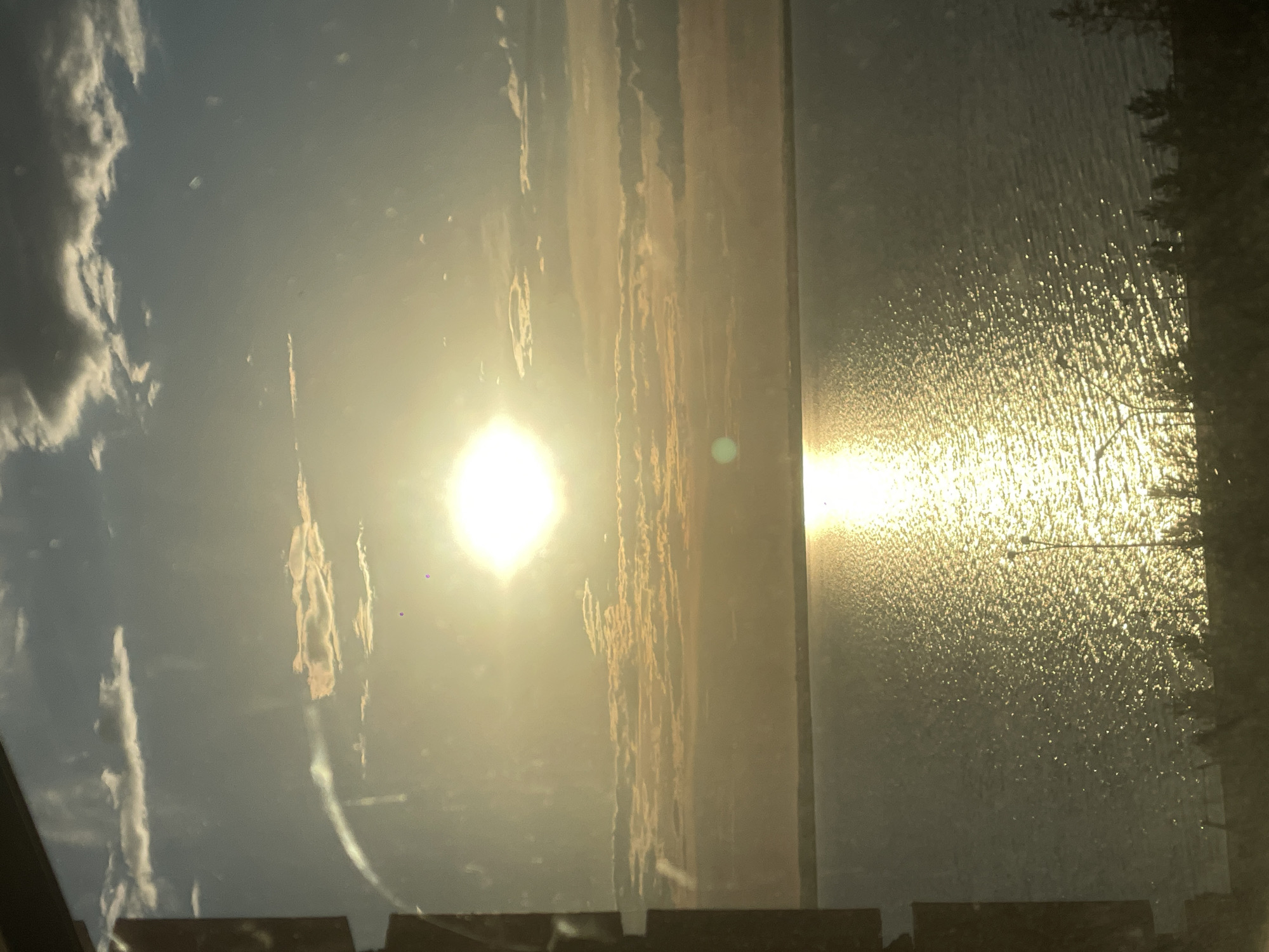 This image depicts a sunset or sunrise over a large body of water, likely Lake Kopa in Kokshetau, Kazakhstan. The sun, a brilliant white-yellow light source with a prominent square-shaped diffraction pattern, is positioned in the upper-left portion of the sky. It projects a bright, shimmering reflection path across the water's surface, extending from the horizon towards the foreground. The sky transitions from a lighter blue in the upper regions, featuring scattered cumulus clouds of white and dark grey, to a warm, hazy golden-orange near the horizon where more stratified cloud formations are visible. The water's surface, particularly outside the direct sun reflection, appears dark blue-grey with visible ripples and small waves. A narrow, dark band of land forms the shoreline in the lower foreground, silhouetted against the sunlit water. This foreground includes indistinct, silhouetted vegetation, possibly small trees or bushes, and the upper portion of a streetlight or utility pole visible on the right. A dark, vertical structure, consistent with a window frame or similar obstruction, is present along the far-left edge of the image. Minor lens flares, appearing as small purple and green dots, are discernable in the upper sky, indicating the photograph was taken through a pane of glass or an optical medium. No people or discernible human activity are present.