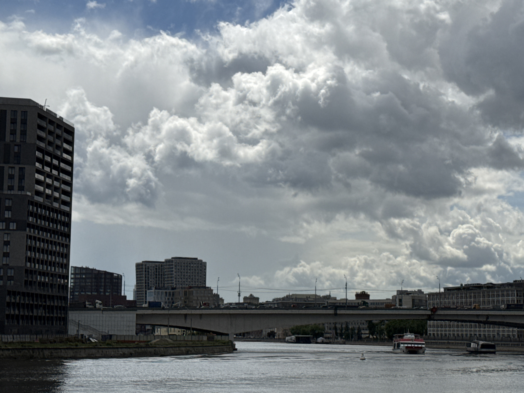The image captures a wide river scene in Moscow, Russia, under a dramatic sky filled with swirling, gray-white cumulus clouds. A large, modern, dark gray building stands prominently on the left, its facade consisting of numerous windows. Further back, a cluster of similar, though smaller, buildings is visible behind a concrete embankment. A concrete bridge spans the river, with vehicles visible on the bridge. In the water, a red and white passenger ferry moves towards the foreground, trailed by a smaller boat. The water itself is gray and choppy, reflecting the overcast sky. A lone white buoy bobs in the water, marking a navigational point. The background is completed by more buildings lining the river, and the bridge.