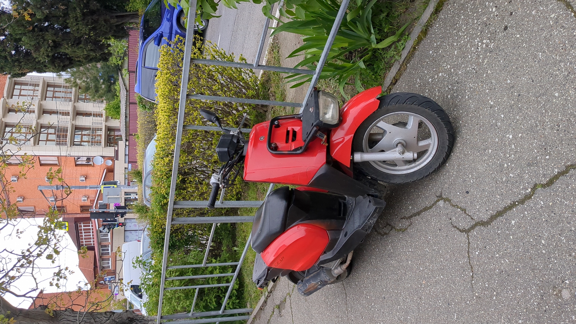 A red Yamaha Gear scooter sits parked on a sidewalk adjacent to a metal fence and greenery. The background includes buildings, trees, and vehicles, suggesting an urban or suburban area. The location is likely Gelendzhik, Russia, based on the scooter model commonly found in the city.