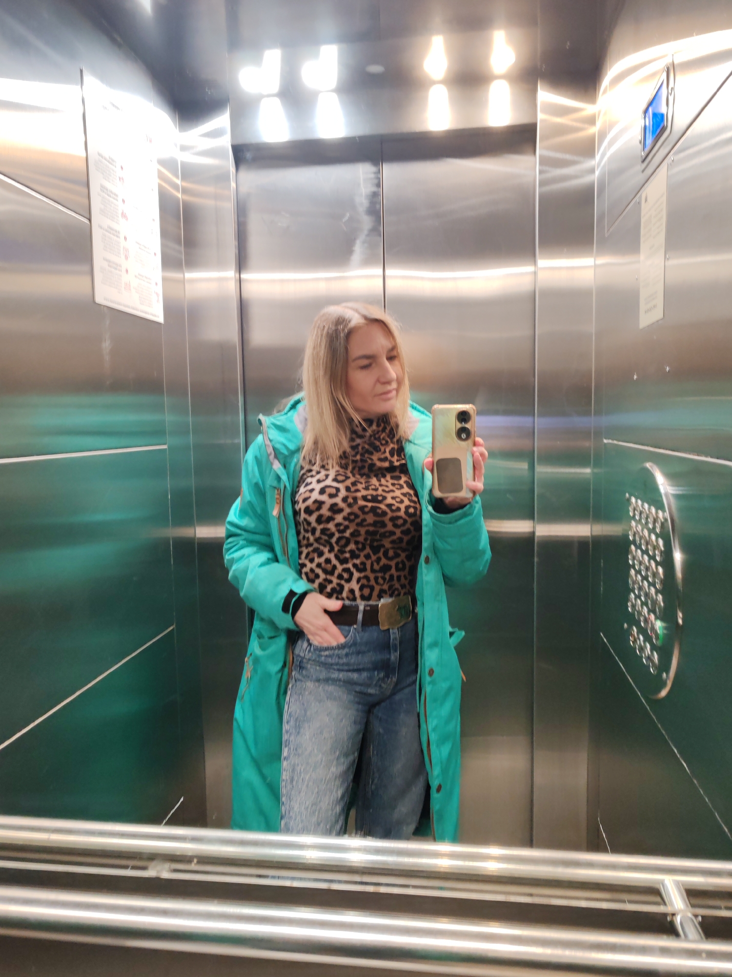 A fair-skinned, blonde-haired woman is seen taking a selfie inside an elevator. She is wearing a turquoise jacket over a leopard print top, and blue jeans with a notable belt buckle. She holds a smartphone with a light green case. The elevator interior is stainless steel, featuring buttons on the right wall and informational signs on the left. The reflection in the bottom of the elevator indicates the presence of metal railings. There is nothing to suggest the location is Saint Petersburg, Russia.