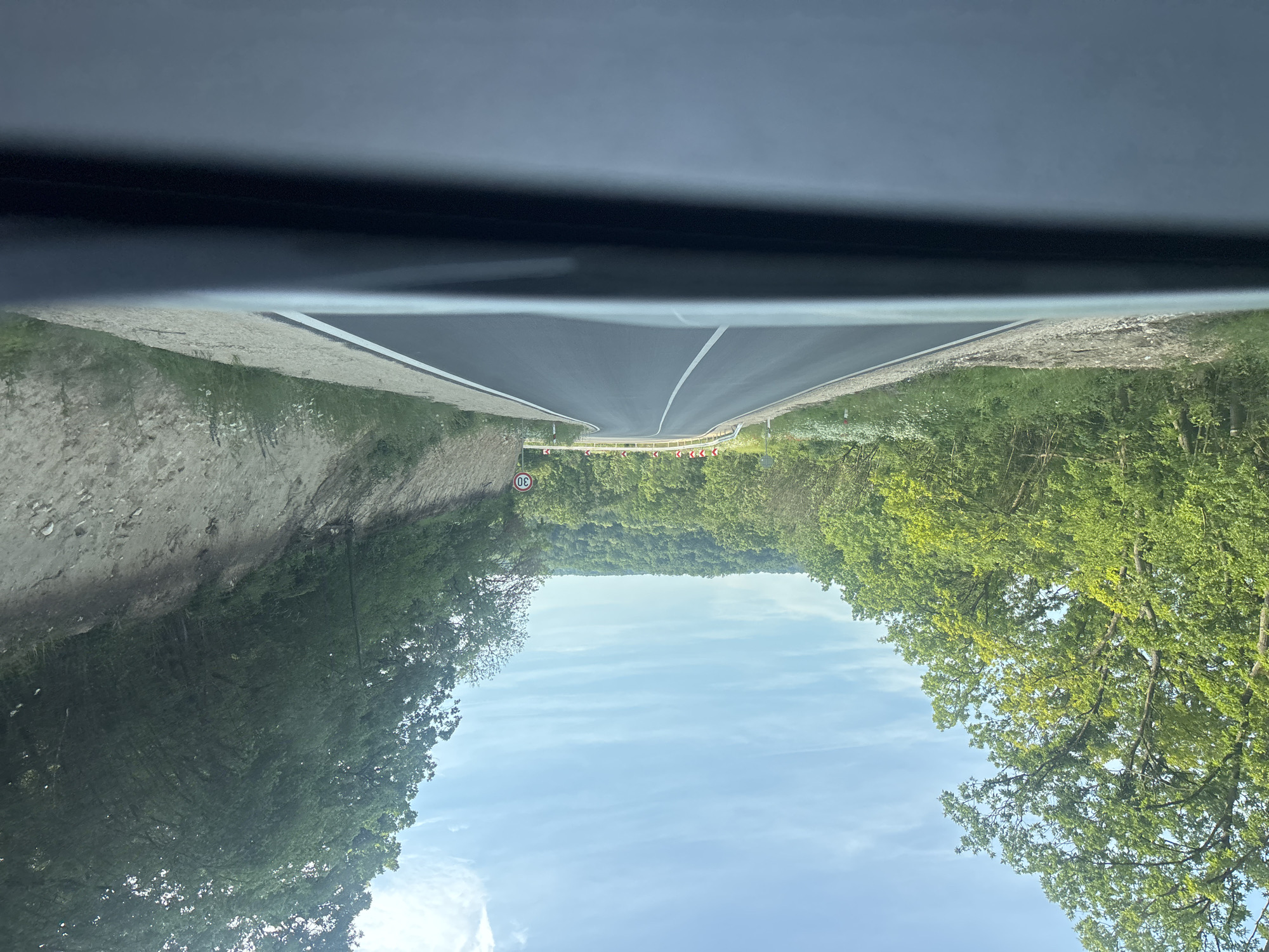 The image depicts a paved asphalt road winding through a densely forested, hilly landscape under a bright, partly cloudy blue sky, captured from the interior of a vehicle looking forward through the windshield. The scene is located in the vicinity of Batovo, Bulgaria.
The road features clear white markings: a solid line defining the right shoulder and a dashed line separating the lanes, indicating a single lane in each direction. The road curves sharply to the left in the mid-ground. Traffic signage is present on the right side of the road, including a circular red sign with a white "30" (denoting a 30 km/h speed limit) and a series of white chevron arrows on red rectangular signs indicating the sharp left turn ahead.
On the left side, dense green deciduous trees and undergrowth border the road, with some small white flowering plants visible on the unpaved shoulder. A red and white reflective marker post is discernible on the left shoulder before the curve. On the right side, the road is bordered by a steep, light brown earthen embankment that transitions into dense green forest. Distant forested hills are visible beneath the horizon.
The vehicle's dark gray dashboard is visible across the bottom of the frame, indicating the perspective. No people, animals, or other vehicles are visible within the frame. The lighting suggests daytime, possibly late morning or early afternoon, with clear visibility. The road appears recently paved or well-maintained.