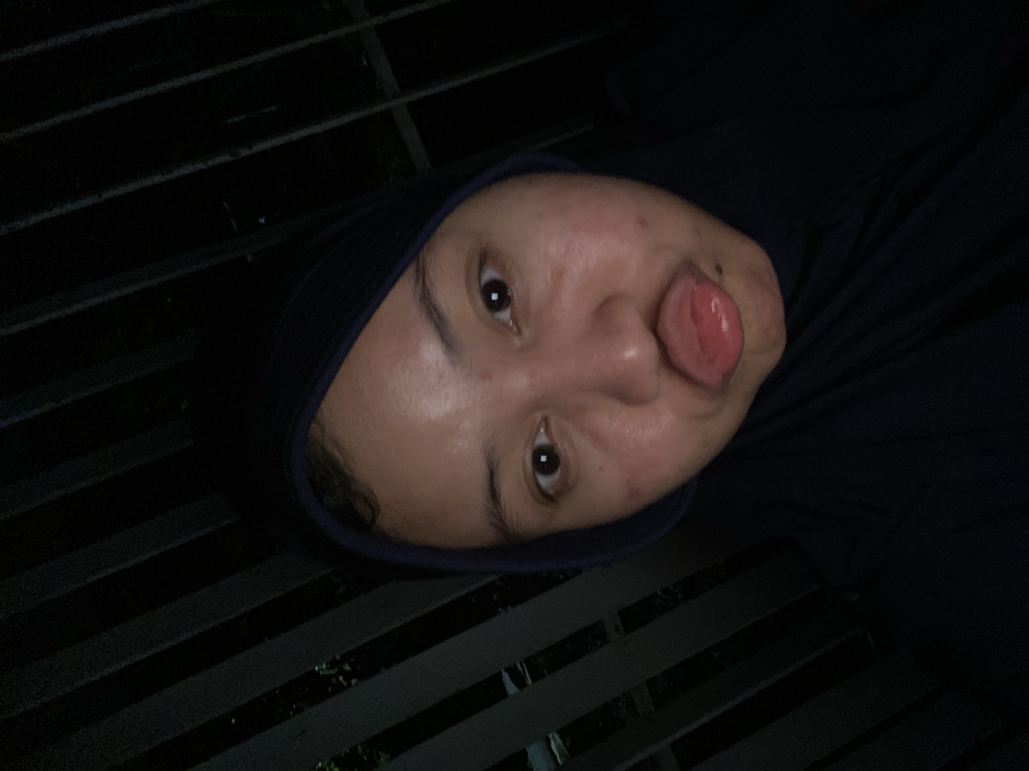 A close-up, selfie-style photograph shows a young woman wearing a dark hijab. She is positioned in the foreground, with her face taking up most of the frame. Her eyes are open and looking towards the camera. Her lips are pursed and her tongue is slightly protruding. The background consists of a dark, out-of-focus area with vertical lines, suggesting a fence or railing. The lighting is dim, and the overall atmosphere is dark. The image does not provide any specific visual cues to identify its location within the city of Banjarbaru, Indonesia.
