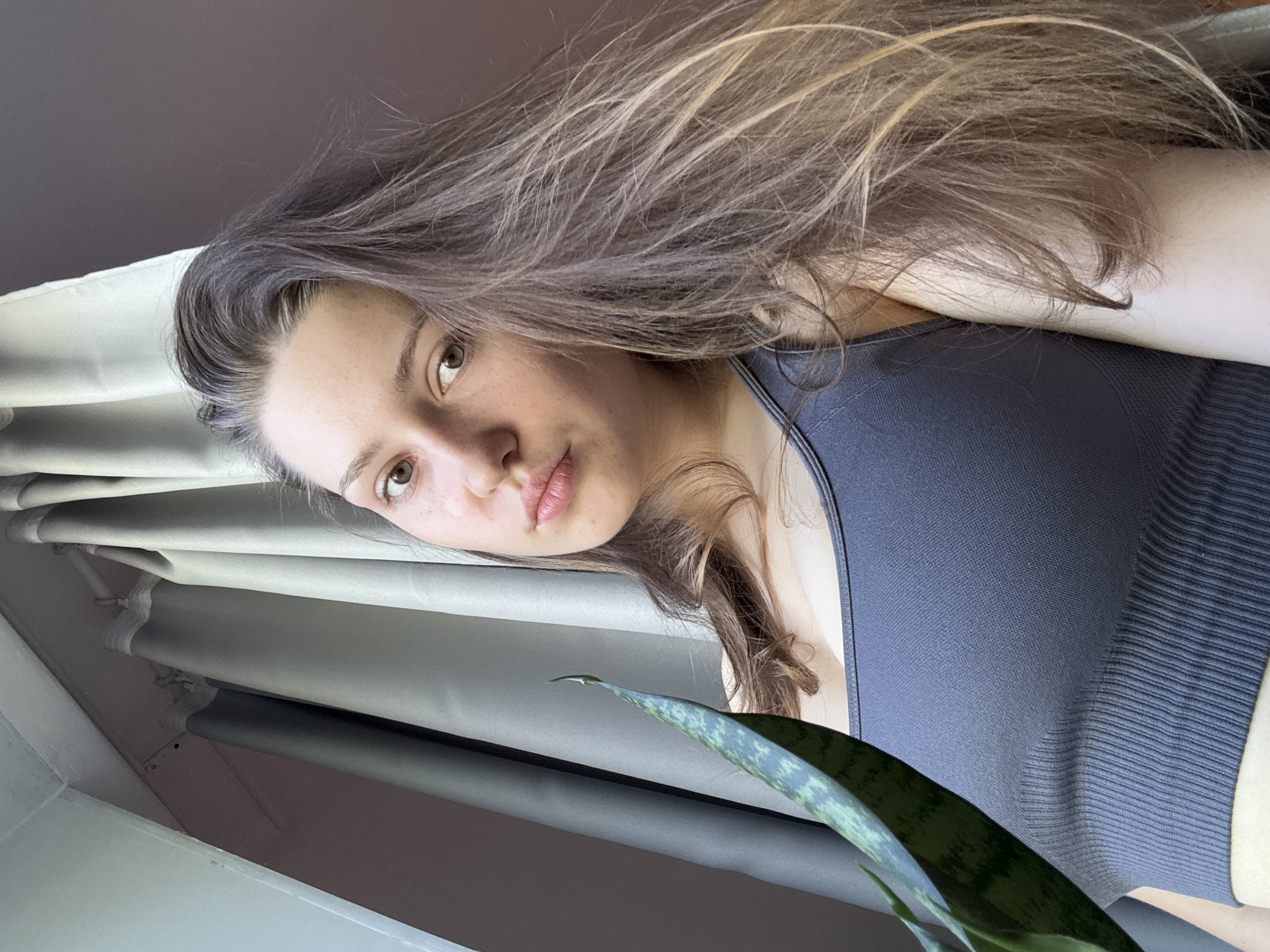 A close-up portrait shows a young woman, with the image rotated approximately 90 degrees clockwise from an upright perspective. She looks directly at the camera with a neutral to slightly pensive expression. Her long, dark brown hair with lighter, possibly blonde, streaks flows around her face and over her shoulders. Her eyes are light-colored, appearing greenish-brown, and her fair skin shows a scattering of freckles. She is wearing a dark grey or blue ribbed sports bra or athletic top.
In the bottom right corner of the frame, a leaf from a snake plant (Sansevieria) is partially visible in the foreground. The background features vertical blinds or curtains, suggesting a window and an indoor setting, likely a room within a residence. Natural daylight illuminates the scene, indicating it is daytime. No text is visible in the image. The depicted location is Saint Petersburg, Russia.