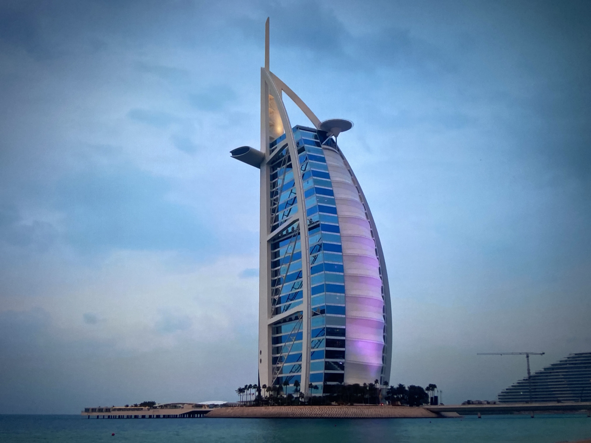 The image depicts the Burj Al Arab hotel in Dubai, United Arab Emirates. It is a sail-shaped building standing on its own artificial island. The sky is cloudy with shades of blue and gray. The water is calm. In the background, another structure is visible, along with a crane. A small line of trees lines the edge of the island.