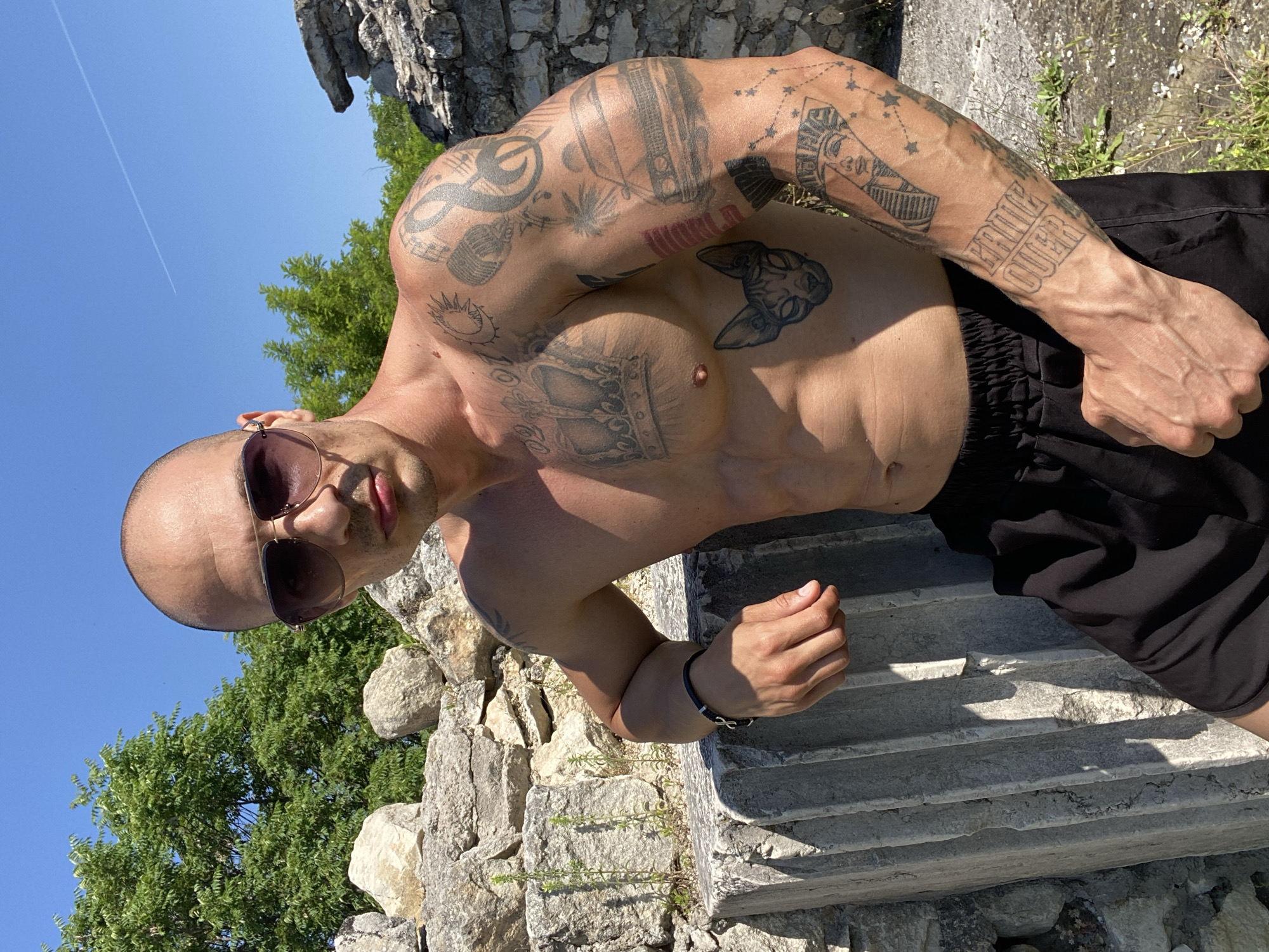 An outdoor, full-body portrait captures a man with a shaved head, wearing sunglasses and black shorts, leaning against a stone structure. He is shirtless, displaying numerous tattoos on his chest and arms, including a crown, a cat, a car, and Egyptian figures. The background features a clear blue sky, green trees, and weathered stone ruins. The scene is illuminated by bright sunlight, casting shadows on the man and the ruins. The man appears to be posing for the photograph. The location is identified as the Pleven Panorama, a historical site in Pleven, Bulgaria, known for its preserved ruins and panoramic views.