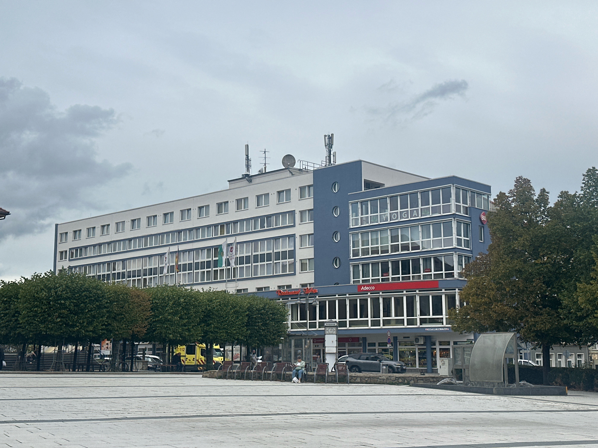 The image shows a modern building in Bautzen, Germany, with a light-colored facade and blue accents. The building features multiple floors with numerous windows, and various antennas are visible on the roof. In front of the building, there is a public square with trees, benches, and a contemporary water feature. Several people are present in the square, some sitting on benches and others walking. A yellow ambulance is parked to the left, and cars are visible on the street behind the trees. The sky is overcast, indicating a cloudy day. Signs for businesses like "Adecco" and "Restaurant Bautzen" are visible on the building's facade.