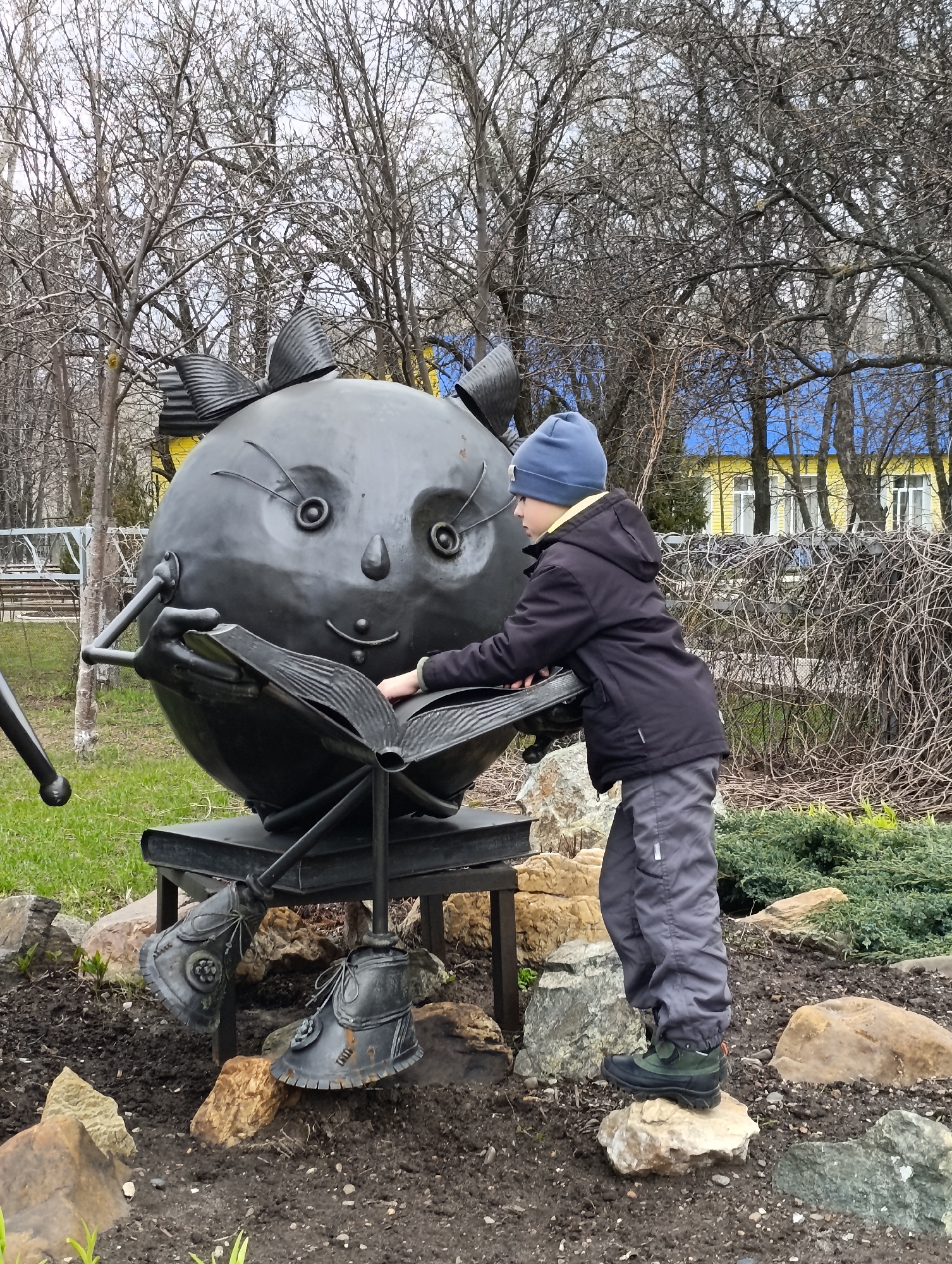 A young boy interacts with a large, spherical metal sculpture in what appears to be a park or public space in Chaikovsky, Russia. The sculpture depicts a whimsical, cartoonish character, possibly a cat, with a round, dark metallic body. It features large, protruding eyes with circular irises, thin antennae-like eyebrows, a small nose, and a simple smile. Decorative elements on its head resemble ears with bows. The sculpture is seated, holding a large, open metal book, and its feet, clad in detailed metal boots, rest on the ground.
The boy, dressed in a blue knitted hat, a black winter jacket, grey insulated pants, and dark boots, stands to the right of the sculpture. He is leaning forward, with his right hand gently touching the pages of the open metal book. He appears engaged and curious, looking towards the book or the sculpture's face.
The setting suggests a cool, possibly early spring or late autumn day, indicated by the numerous deciduous trees in the background with bare, leafless branches. The ground around the sculpture is a mix of bare earth, scattered rocks, and patches of green grass and low-lying shrubs. In the background, behind the trees, a building with yellow walls and a bright blue roof is visible. A fence or overgrown hedge with intertwined brown branches runs behind the boy. The sky is overcast. No discernible text is visible on any of the subjects or in the background.