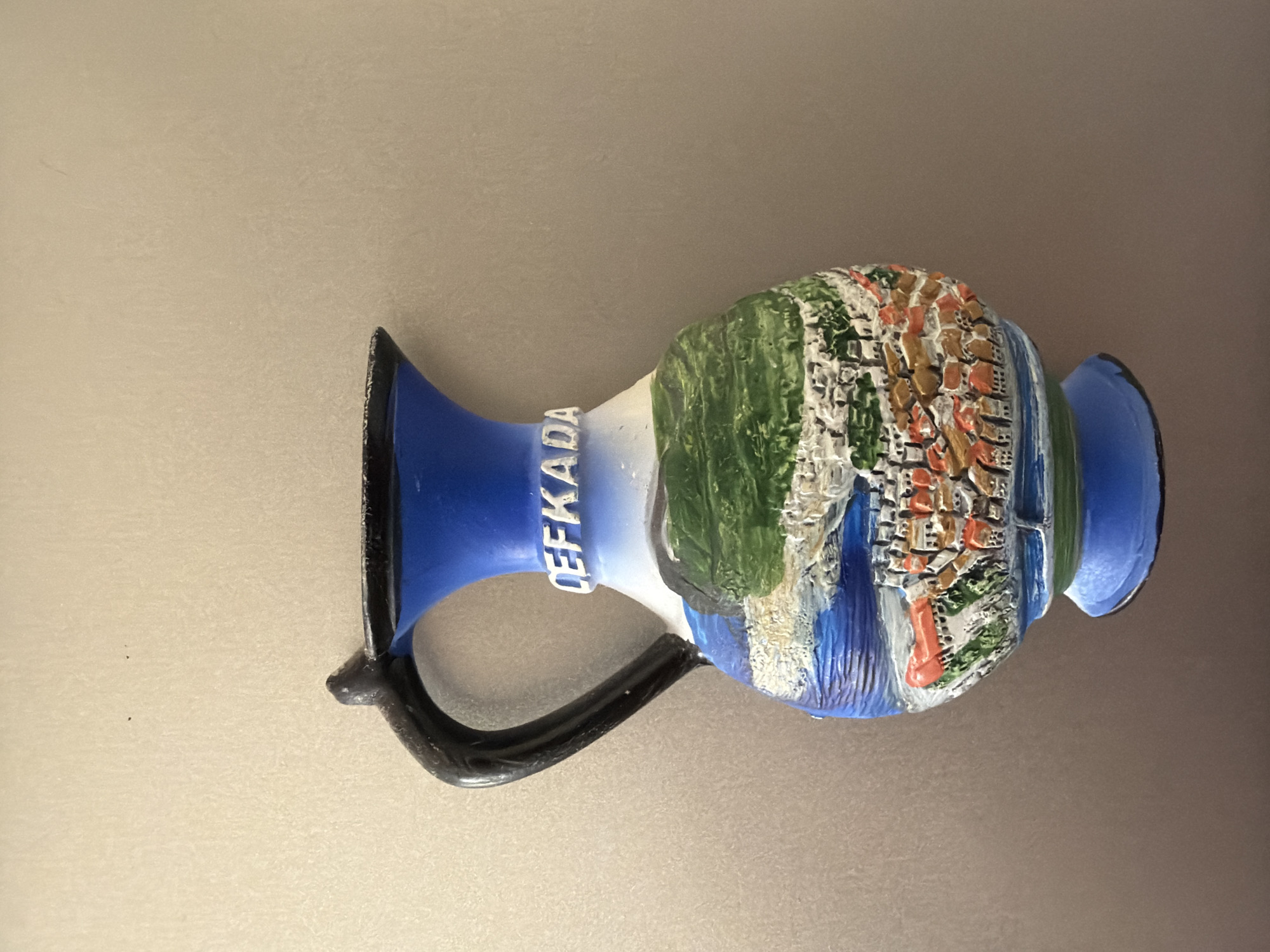 A souvenir vase is displayed against a plain, textured, beige-grey background. The vase is shaped like a jug with a handle. The top of the vase is blue with a black rim. White letters spelling "CEFKADA" are embossed around the neck of the vase. The body of the vase depicts a raised relief of a coastal town with buildings, green hills, and blue water. The town has red-tiled roofs and appears to be situated on a hillside overlooking the sea. The base of the vase is blue with a green band. There are no people or any indication of the city of Pazardzhik, Bulgaria in the image.