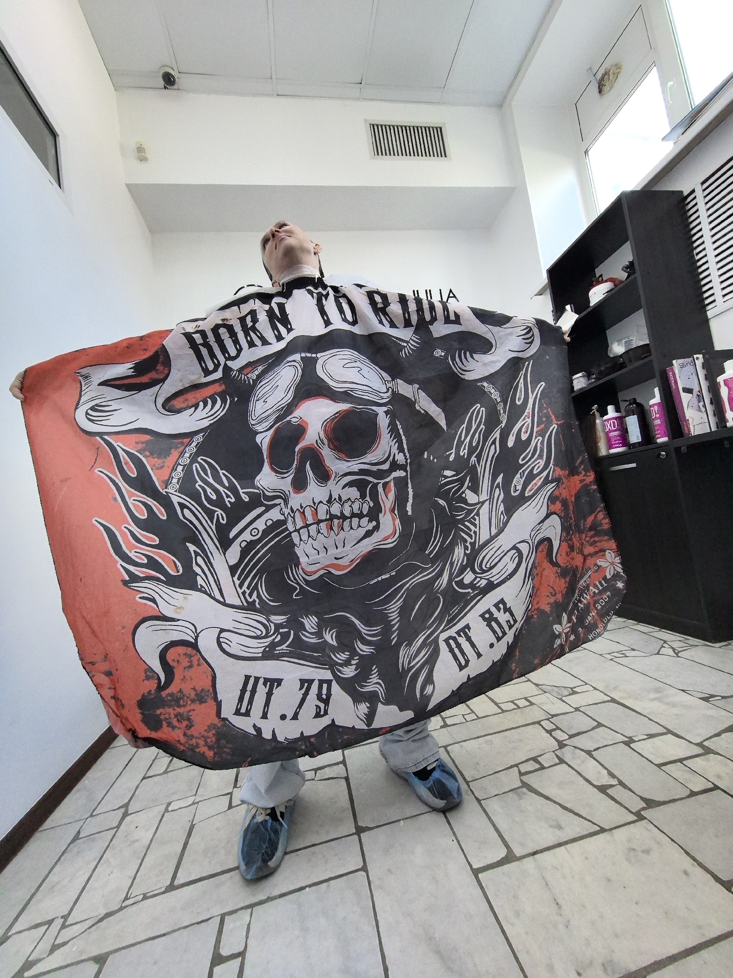 A low-angle, wide shot captures a person standing in what appears to be a barber shop, holding a large cape featuring a skull graphic and the text "BORN TO RIDE." The cape is predominantly red and black, with the skull wearing goggles and surrounded by flames. The person is wearing light-colored pants and blue slip-on shoes. The environment includes a white tiled floor, white walls, a ceiling with lights and a vent, and a black shelving unit filled with various bottles and products. The lighting is bright, coming from above and a window on the right. The composition emphasizes the cape and the person's pose, with the background details serving to contextualize the scene. The image appears to be taken indoors, in a commercial establishment.
