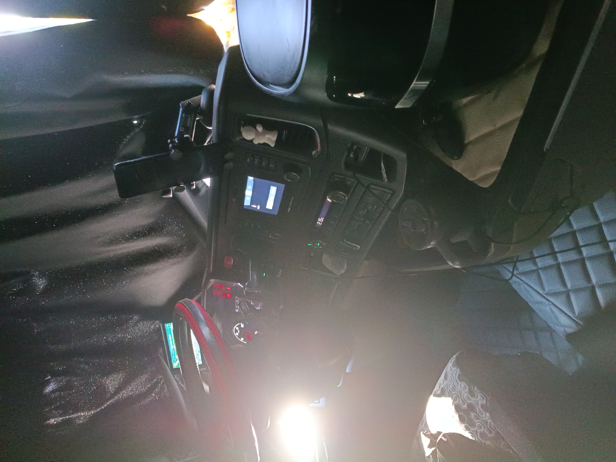 The image shows the interior of a truck cabin, with a focus on the dashboard and driver's seat area. A bright light source, possibly sunlight, illuminates the left side of the image, casting strong glare on the steering wheel and dashboard. The dashboard features various instruments and screens, including a navigation display and a radio. A phone is mounted on a holder on the right side of the dashboard. A small white figurine is visible on the dashboard, near an air vent. The driver's seat on the left is partially visible, with a patterned fabric covering. The floor is covered with quilted mats. The overall environment appears to be indoors, within a vehicle. The location is identified as Uttrichshausen, Germany.