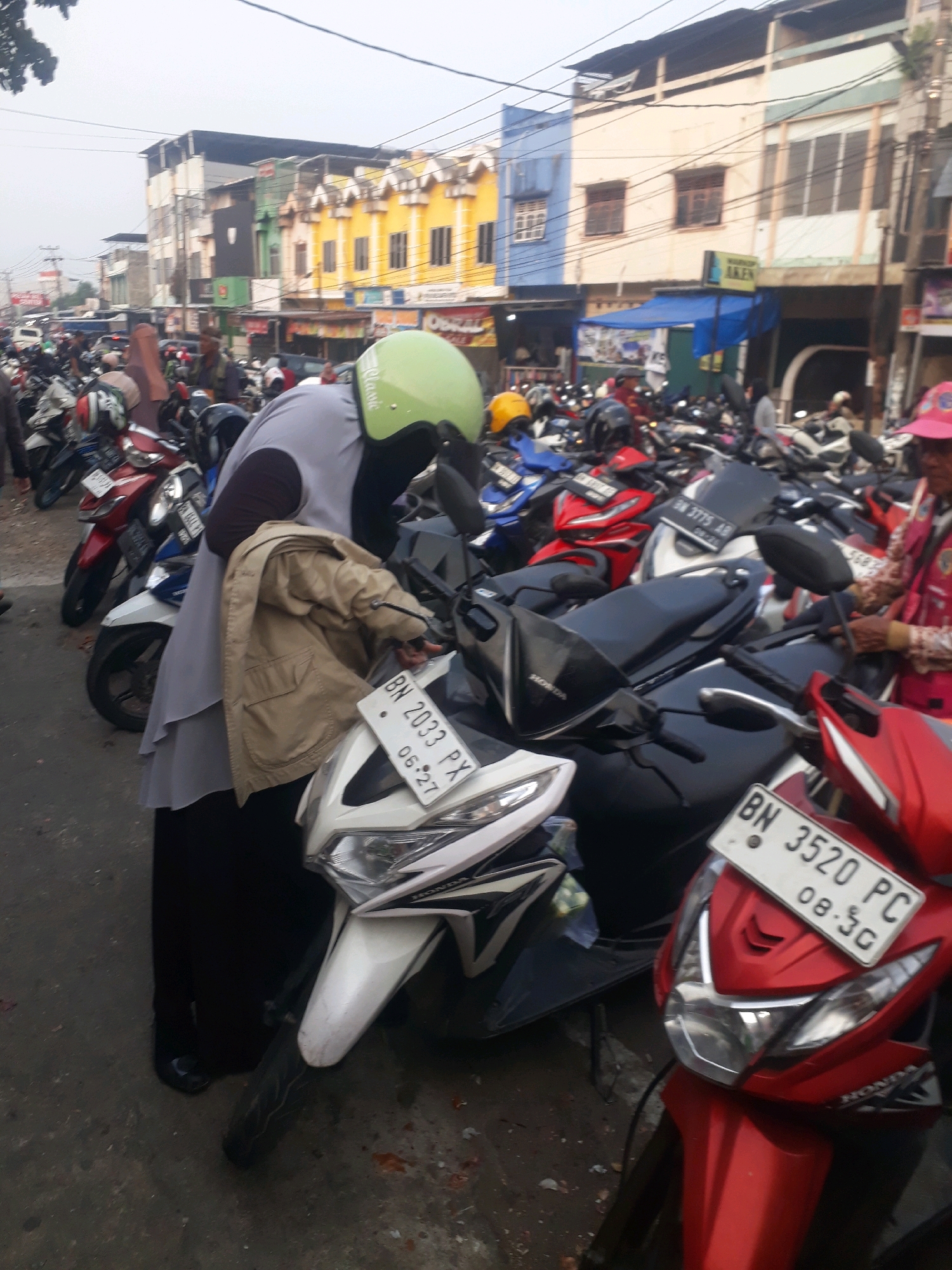 The image depicts a street scene in Pangkalpinang, Indonesia, characterized by a dense parking lot filled with motorcycles. In the foreground, a woman wearing a gray hijab and a light brown jacket is standing next to a white and black motorcycle. Another motorcycle, red in color, is parked to the right, with a license plate visible reading "BN 3520 PC 08.5C". Several other motorcycles, in various colors including red, blue, and white, are packed closely together in the background, suggesting a busy area, possibly near a market or commercial establishment. The buildings lining the street are a mix of colorful and neutral facades, with awnings and signage indicating businesses. The sky is overcast, contributing to the muted lighting of the scene. The overall impression is one of a typical urban environment in Indonesia, with a strong emphasis on motorcycle transportation.