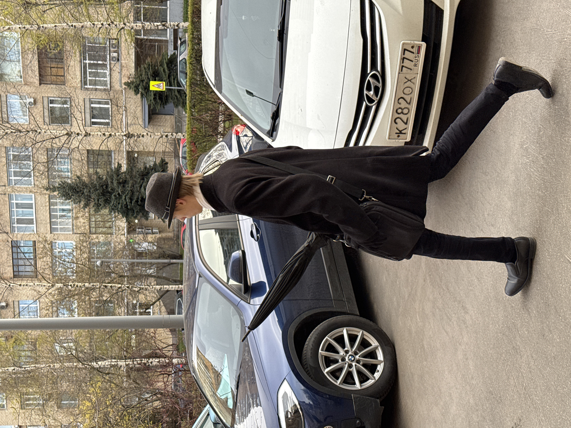 A person wearing a dark coat and hat is walking on a city street. The person is between two cars, a blue one and a white one, parked next to each other. The white car has a Russian license plate with the characters "K282OX777". The person carries a black umbrella and a shoulder bag. The background includes an apartment building and trees.