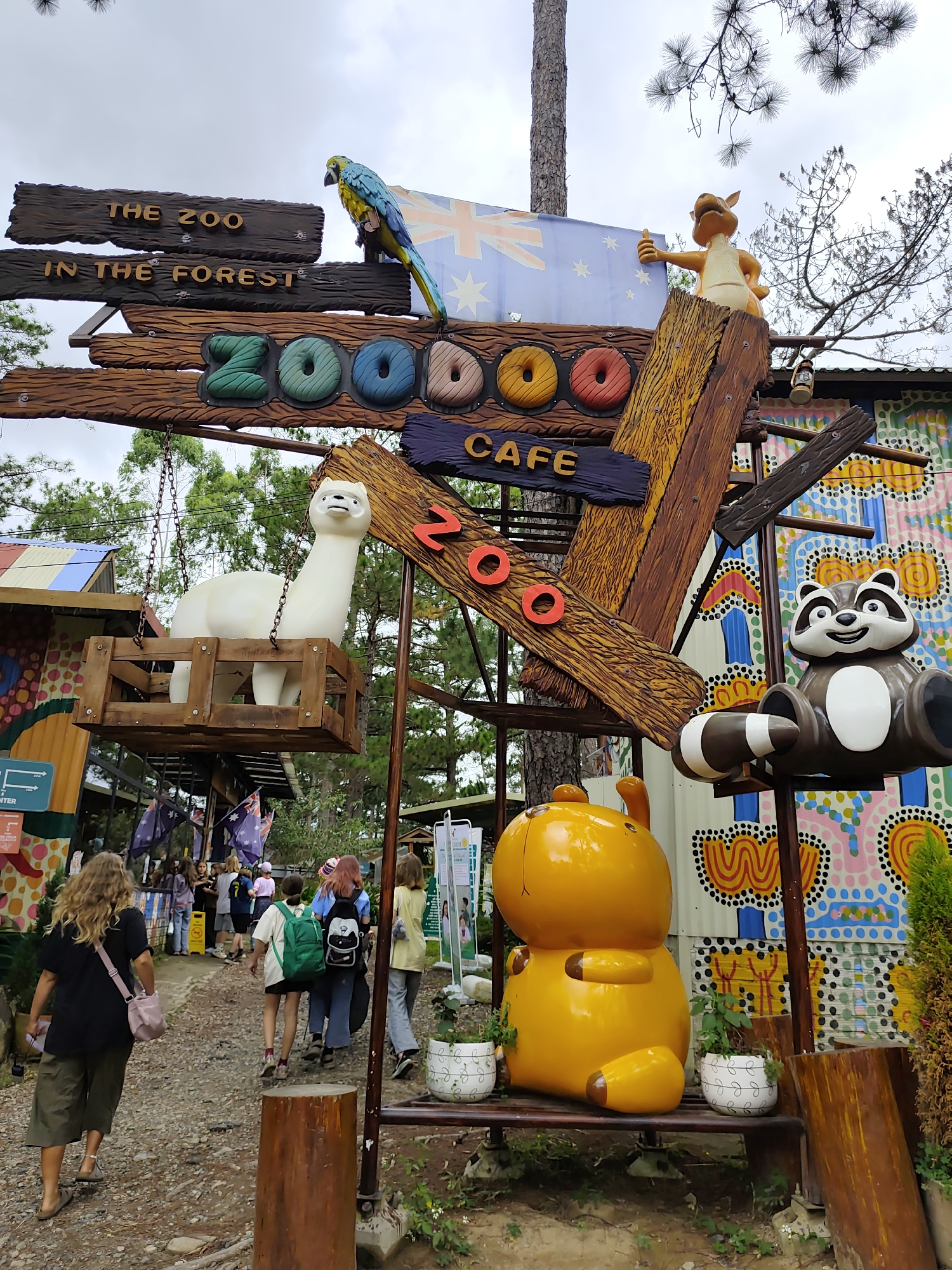 The image shows a whimsical outdoor decoration at a zoo cafe. The central structure is a wooden sign with the words "THE ZOO IN THE FOREST" and "ZOODO CAFE" written on it. The letters for "ZOODO" are shaped like donuts and are colored green, blue, and pink. A bright blue and yellow macaw perches on top of the "ZOO" sign. To the right, a cartoonish kangaroo figure gives a thumbs-up. Below the main sign, a white llama statue is suspended in a wooden crate. To the right of the llama, a cartoon raccoon statue is seated, and a large, shiny yellow cartoon animal figure is sitting on a bench. The background features a colorful, patterned wall. Several people are walking on a dirt path in front of the decorations. The environment appears to be a zoo or an amusement park.