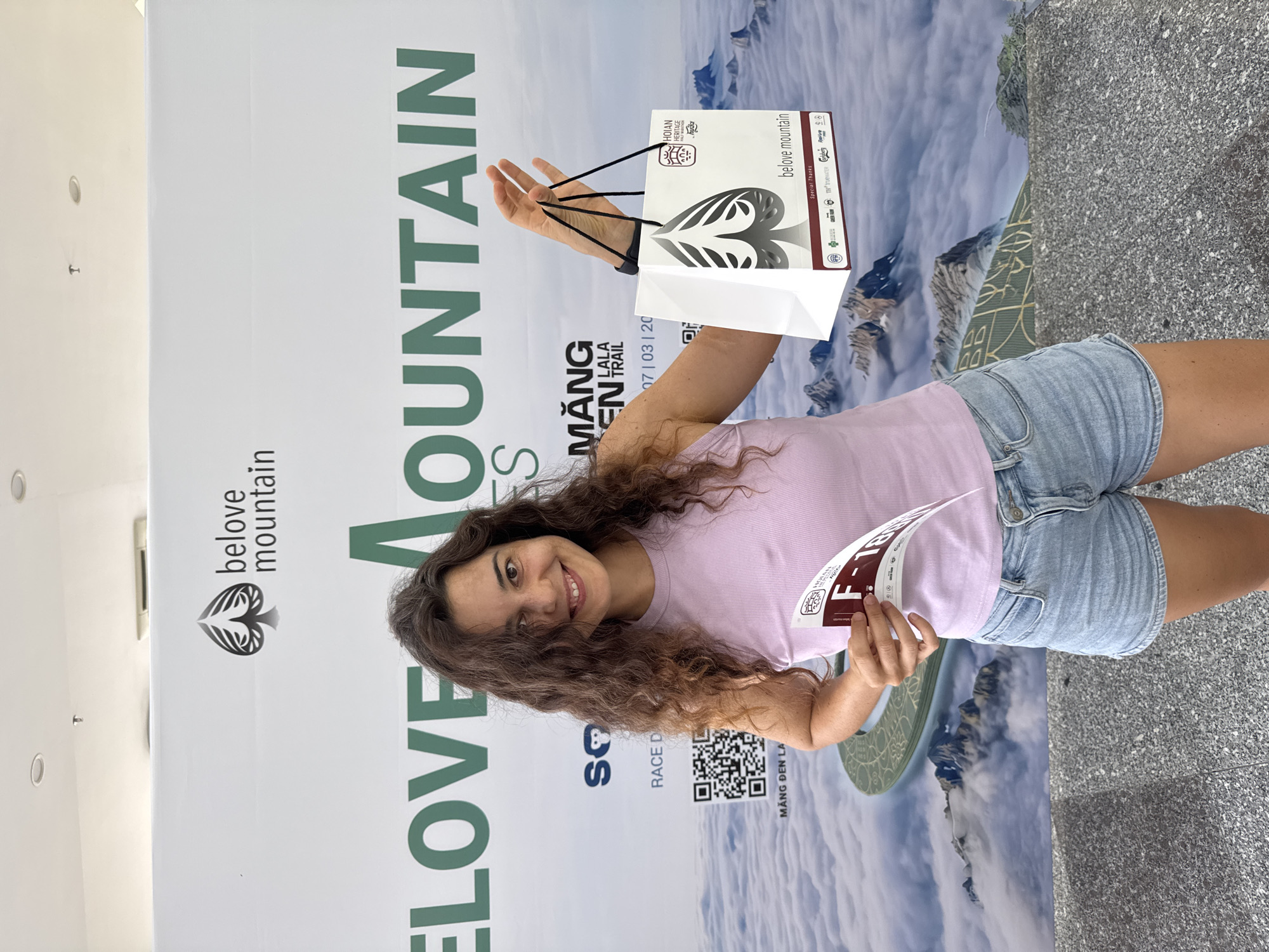 A young woman with long, curly brown hair smiles and holds up a white gift bag and a race bib in front of a large banner. The banner features the text "belove mountain" and "BELOVE MOUNTAIN" in green letters, along with a spade symbol. The gift bag has the same spade symbol and "belove mountain" text, along with "HOI AN HERITAGE RACE" and sponsor logos. The race bib is white with red text that includes "F-188". The background of the banner shows a mountainous landscape covered in clouds. The woman is wearing a light purple tank top and denim shorts. The scene appears to be outdoors, possibly at an event related to the Hoi An Heritage Race.