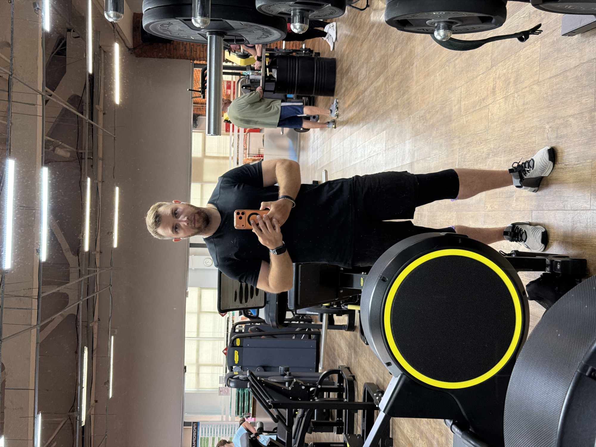 A man stands in a gym, taking a selfie in a mirror. He is wearing a black t-shirt and black shorts, with a black watch on his left wrist and a black bracelet on his right. He is holding an orange-cased smartphone in his right hand. The gym has wooden floors and exposed ceilings with fluorescent lighting. Various weightlifting equipment, including weight plates and machines, are visible in the background. Two other individuals are present in the background, one wearing a green shirt and blue shorts, and another wearing a black top and black leggings. The scene appears to be set in a gym in the city of Chaikovsky, Russia.