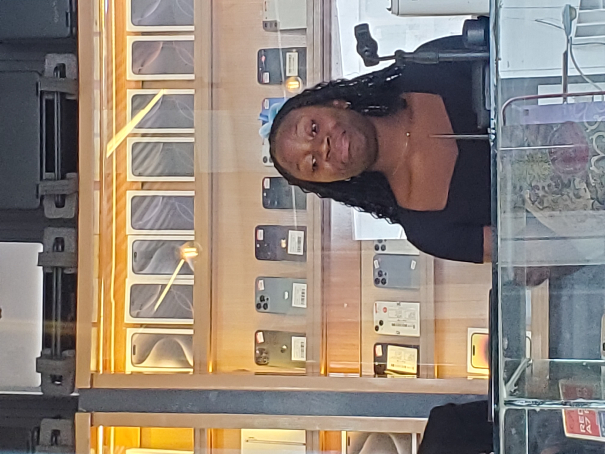 In Jos, Nigeria, a Black woman with braided hair is captured in a photograph, her face turned slightly towards the camera with a neutral expression. She is wearing a black top. The background reveals a retail display of mobile phones, predominantly iPhones, arranged in a grid within a glass case. The phones are presented in their boxes, showcasing various models and colors. The lighting suggests it is daytime, possibly indoors in a well-lit store. Some reflections are visible on the glass, hinting at the surrounding environment. The overall scene depicts a commercial setting, likely a phone shop.