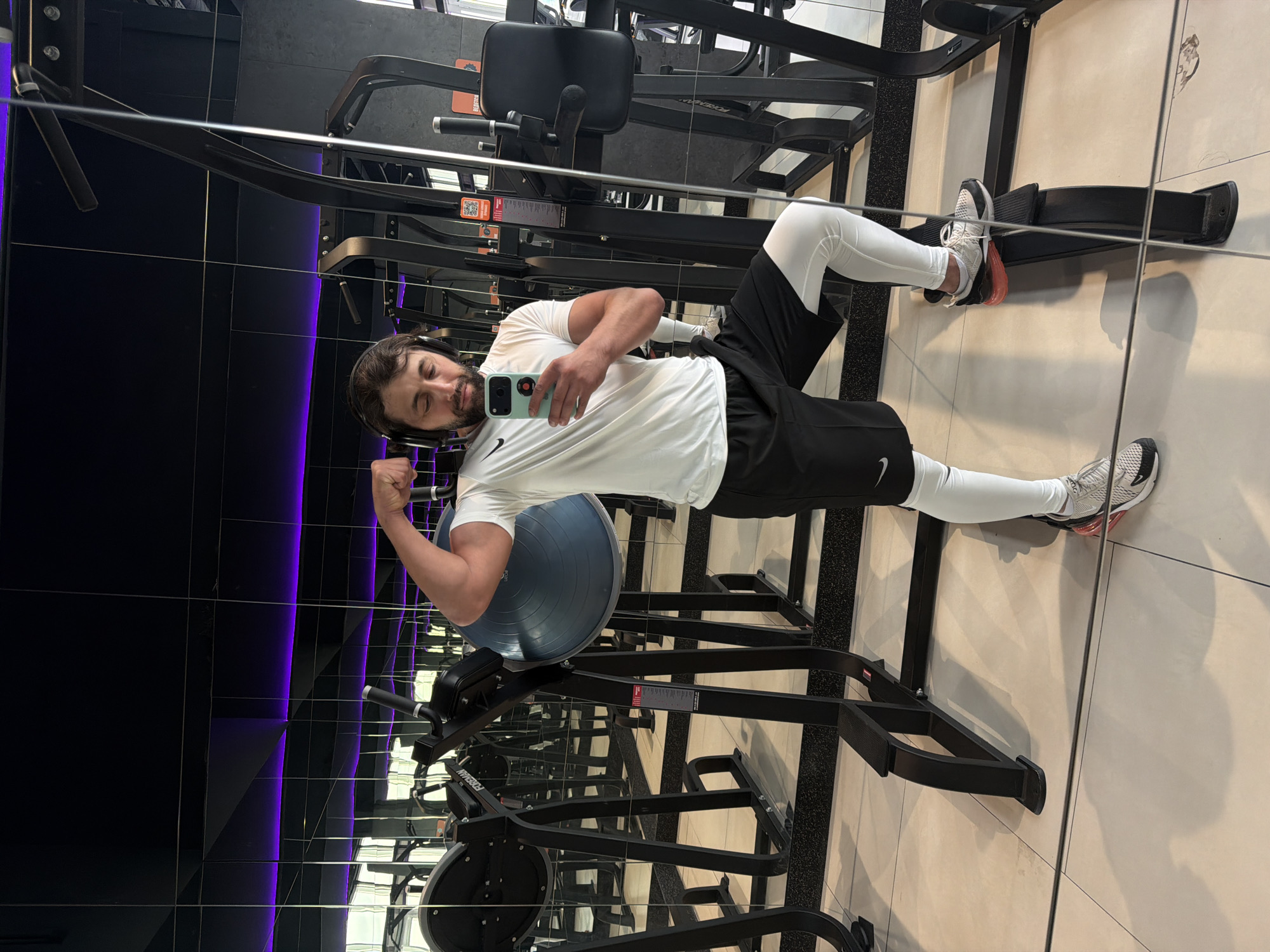 The image shows a man taking a selfie in a gym, likely within the city of Krasnoyarsk, Russia. The man is wearing headphones, a white t-shirt, black shorts, and white leggings with athletic shoes. He is standing next to a piece of exercise equipment and flexing his bicep. The gym has mirrored walls and tiled floors. Other exercise machines are visible in the background.