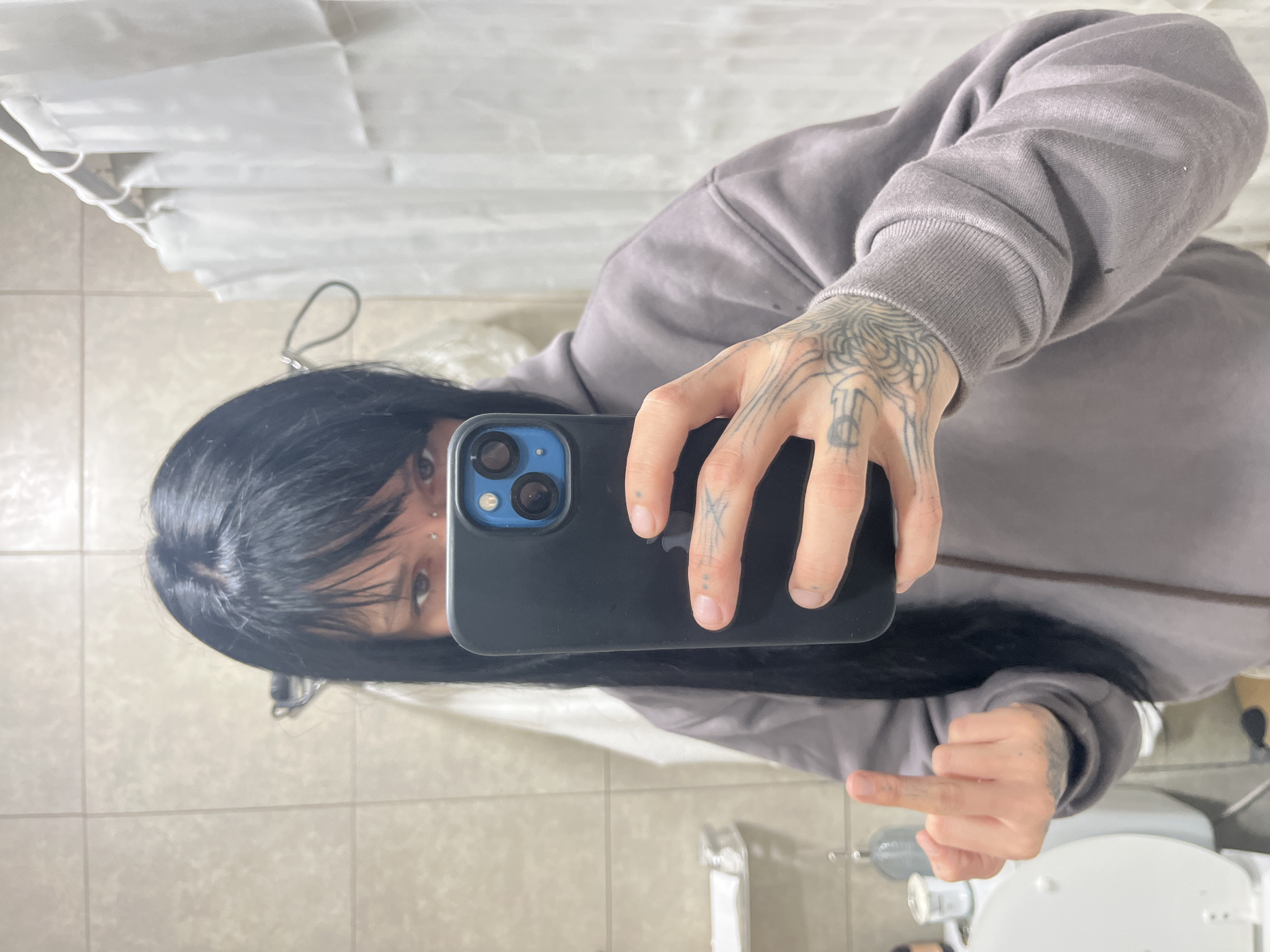A person with long black hair is holding a phone in front of a mirror, taking a selfie. The person is wearing a gray sweatshirt and has visible tattoos on their hands. The person is extending their middle finger. The background includes a bathroom with a tiled floor, a shower curtain, and a toilet. The location cannot be determined from the image.