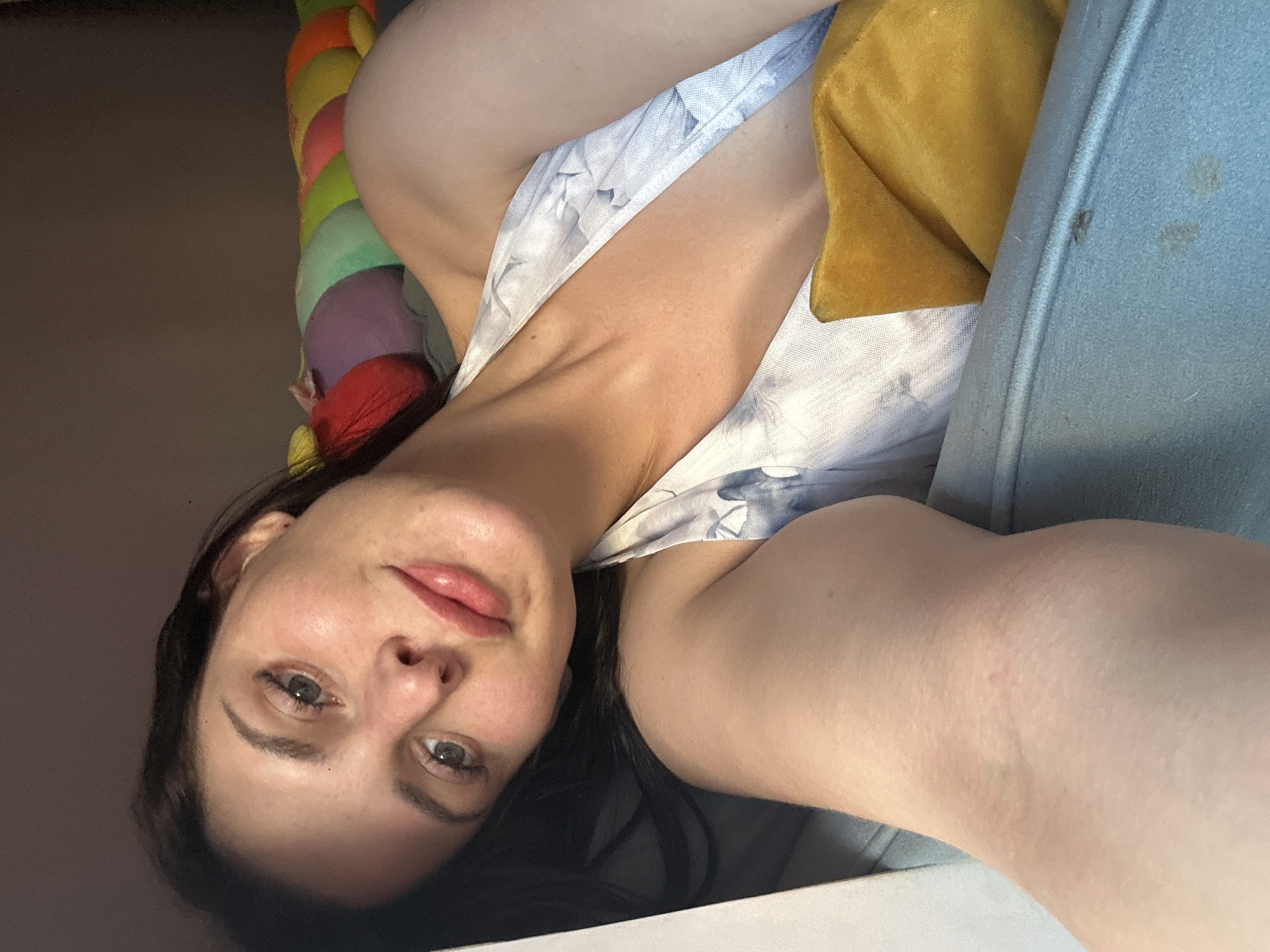 A young woman with dark hair is lying on her side, looking up at the camera. She is wearing a white halter top with a light blue floral pattern. Her skin is fair and she has rosy cheeks and full lips. Her eyes are green and appear to be looking directly into the camera. She is lying on a blue couch, with a yellow pillow tucked behind her head. In the background, there is a colorful, segmented stuffed toy that resembles a caterpillar. The lighting suggests the photo was taken indoors, possibly in a bedroom or living room.
